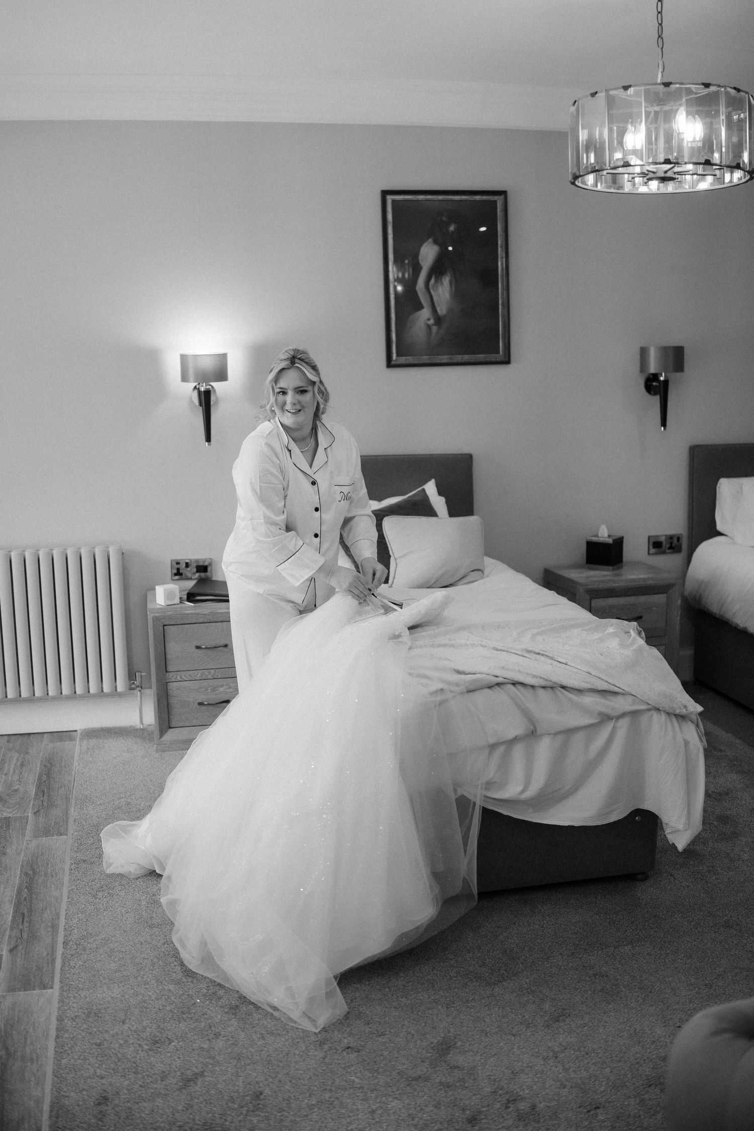 Bride in wedding dress and pyjama top sitting on hotel bed getting ready in black and white photo.