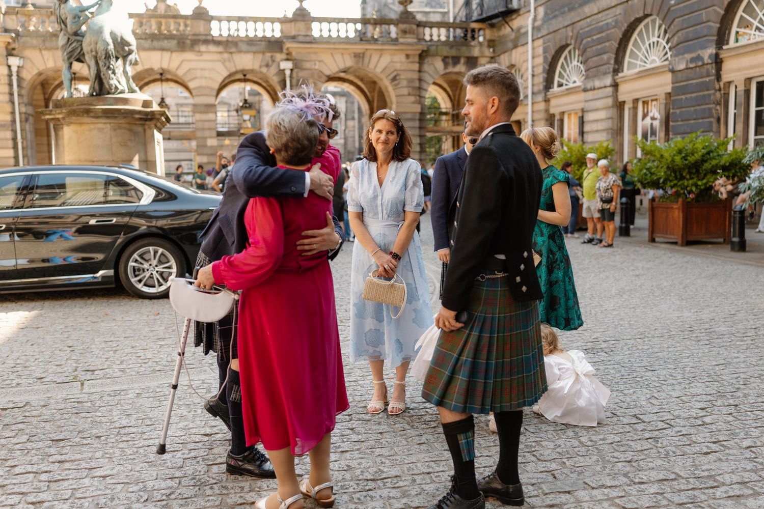People in formal attire, including a man in a kilt, greet each other outside a grand stone building.