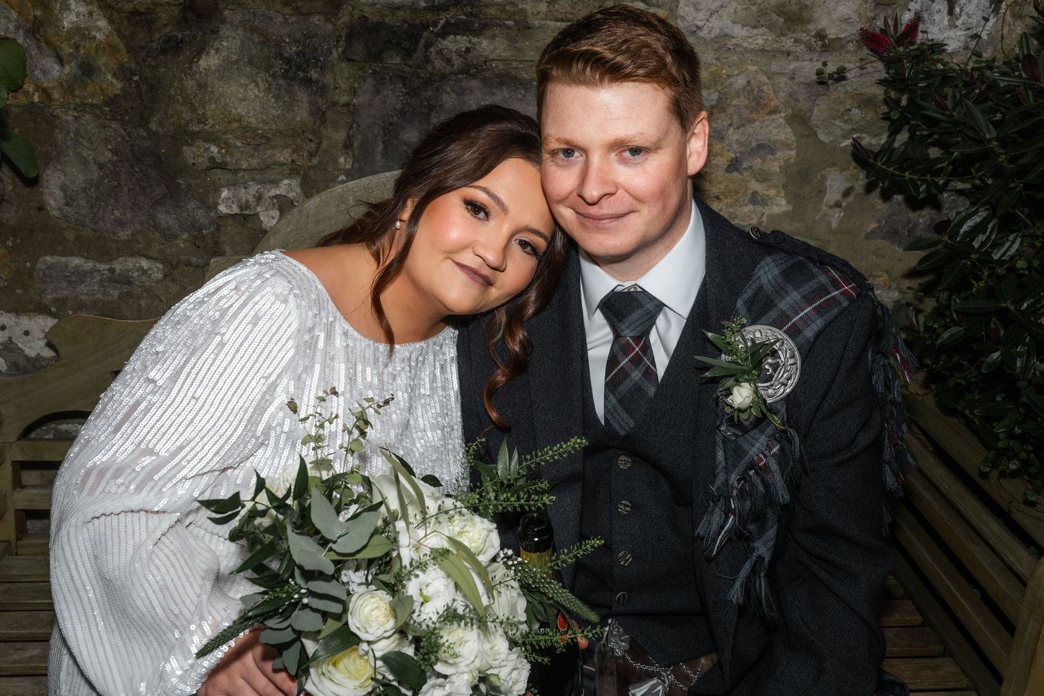A couple in wedding attire poses together, with the bride holding a white and green bouquet.