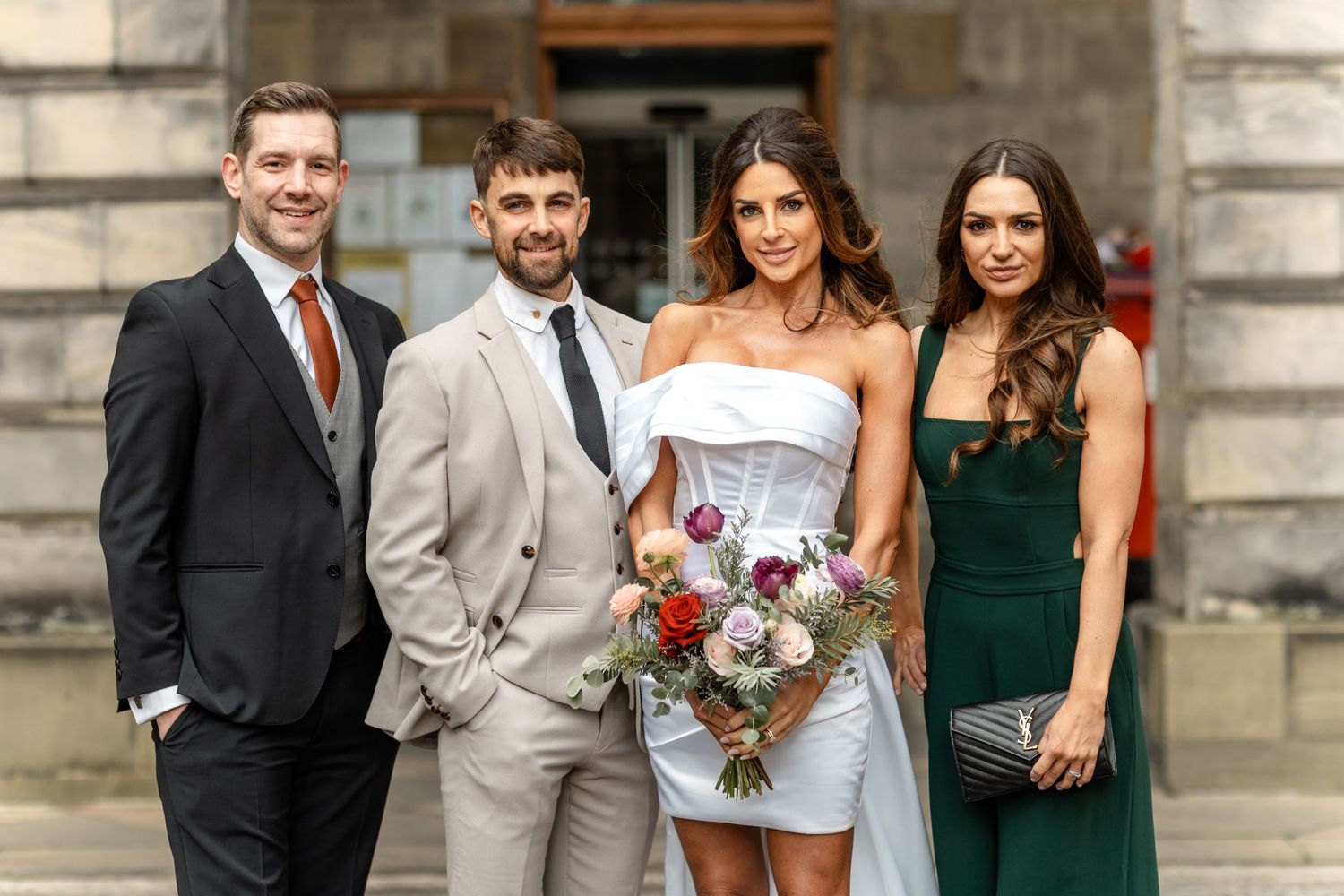 Wedding party poses in front of historic stone building with bride in short white dress.