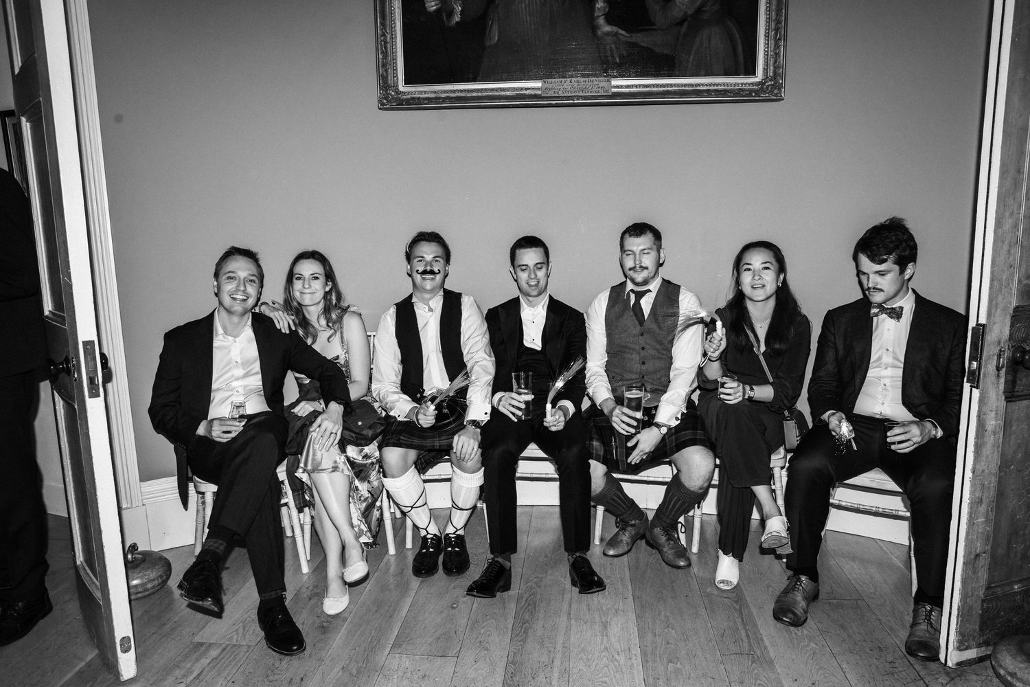 Black and white photo of wedding guests sitting along a wall at a reception venue.