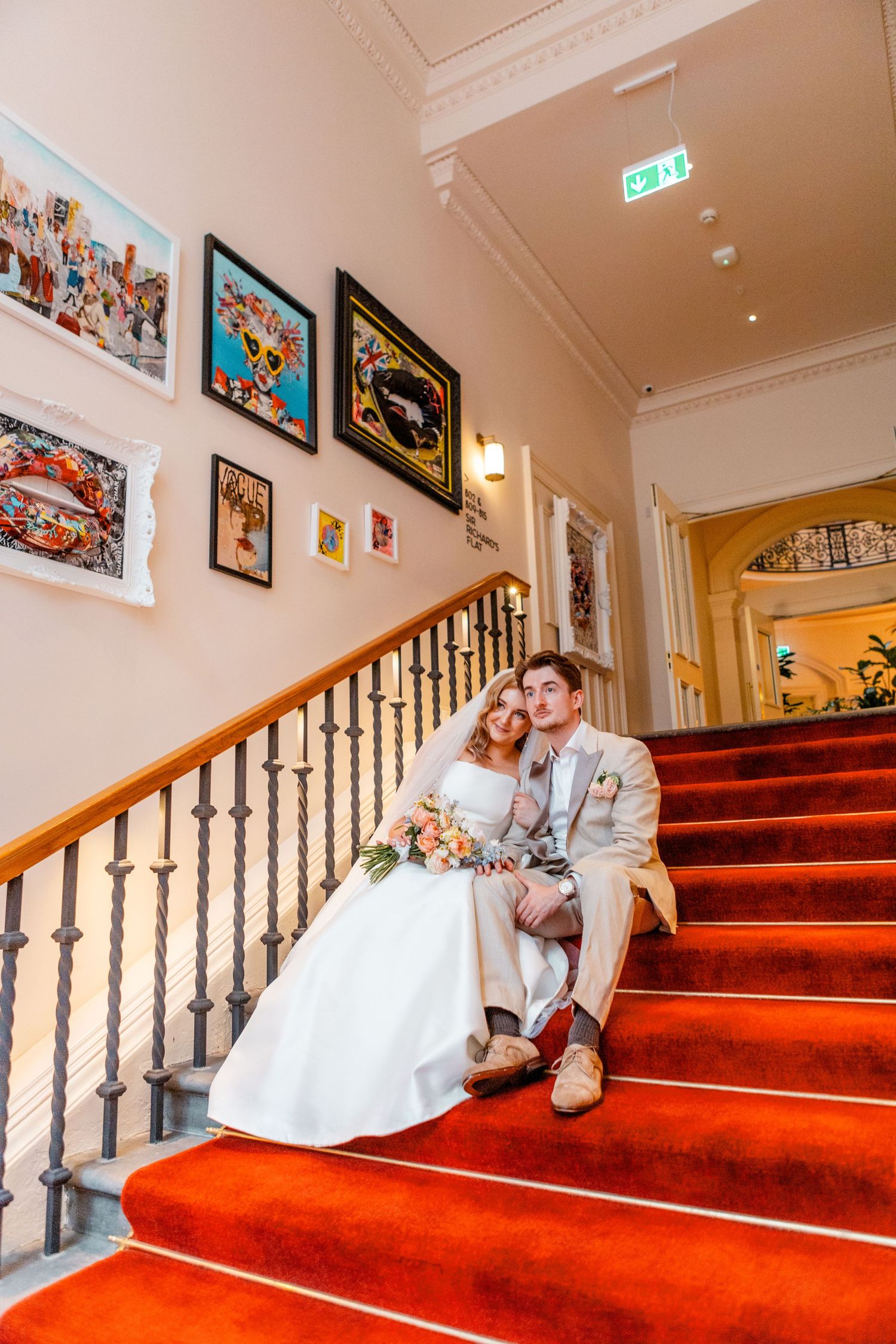 Wedding couple relaxes on red carpeted staircase decorated with framed artwork.