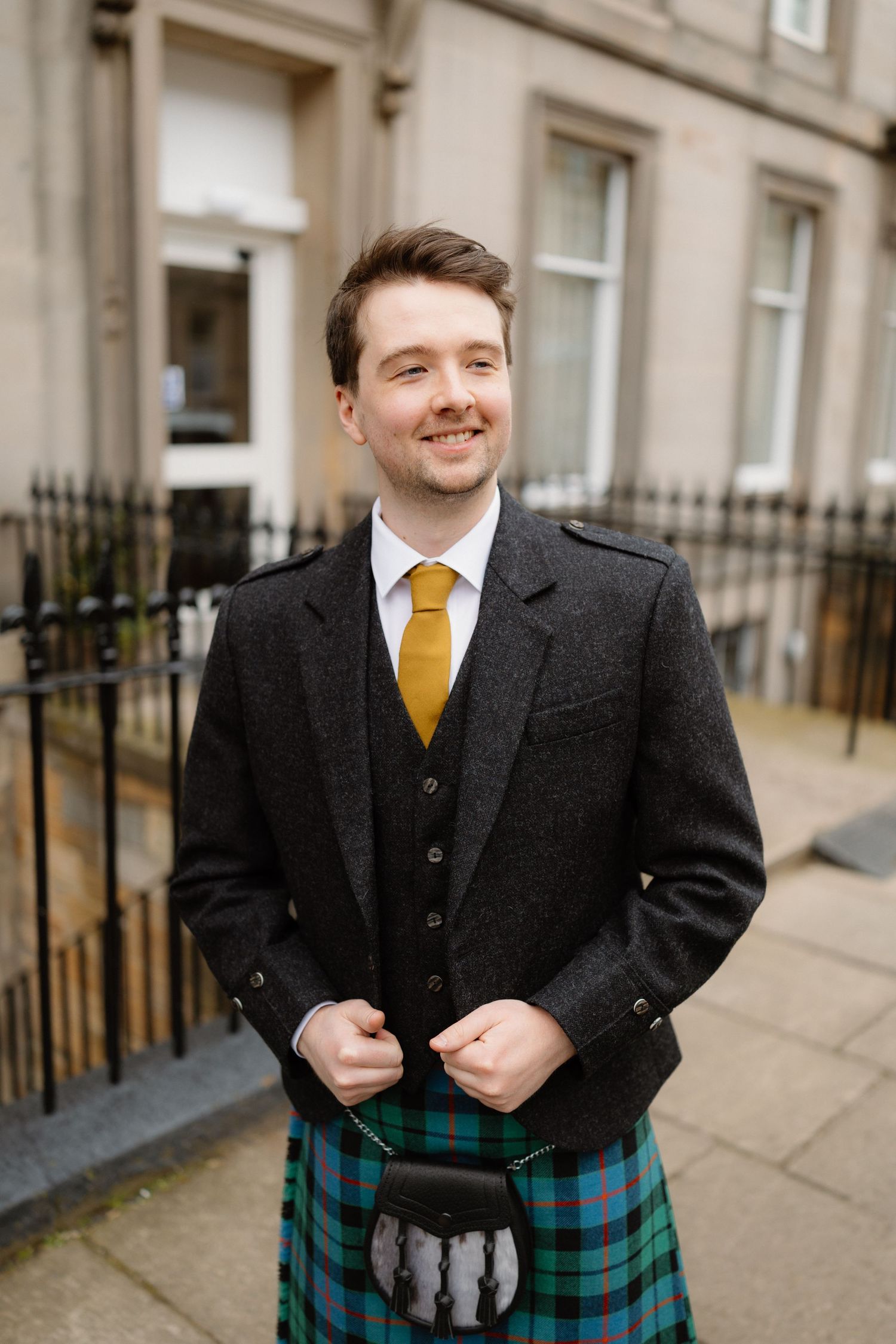 Smiling groom in traditional Scottish kilt and charcoal jacket with gold tie standing on a city street.