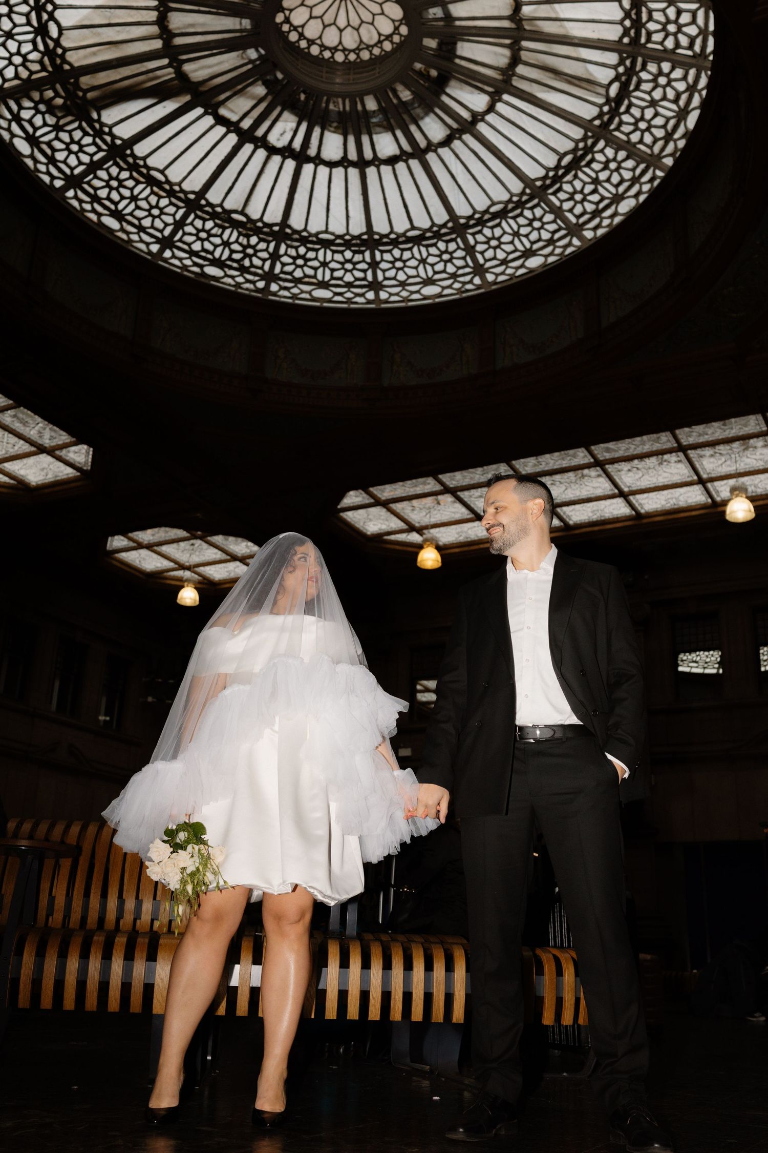 Bride in short white dress and veil holds bouquet with groom under ornate glass dome ceiling.