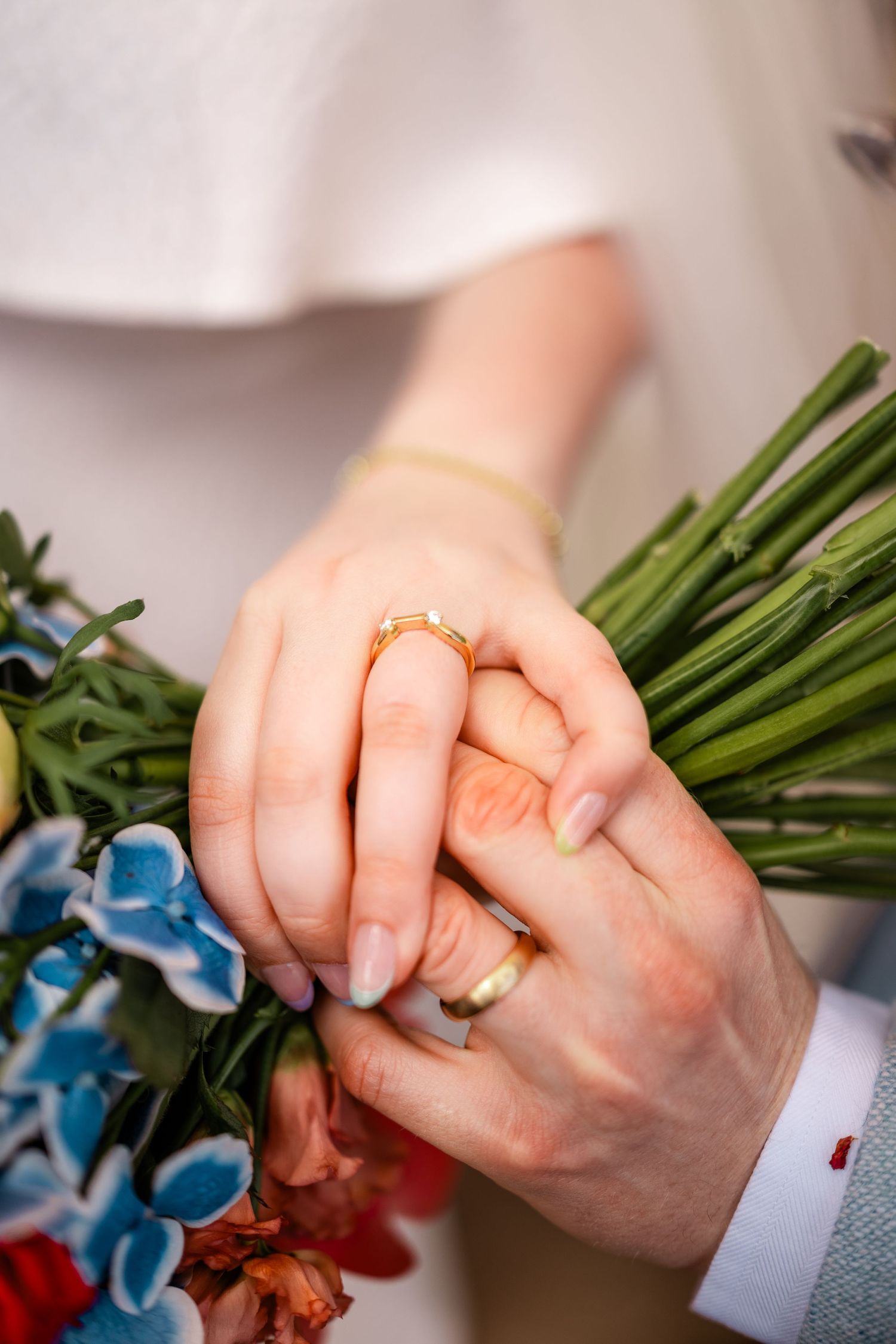 Close-up of newlyweds holding hands with gold wedding rings over colorful bridal bouquet.