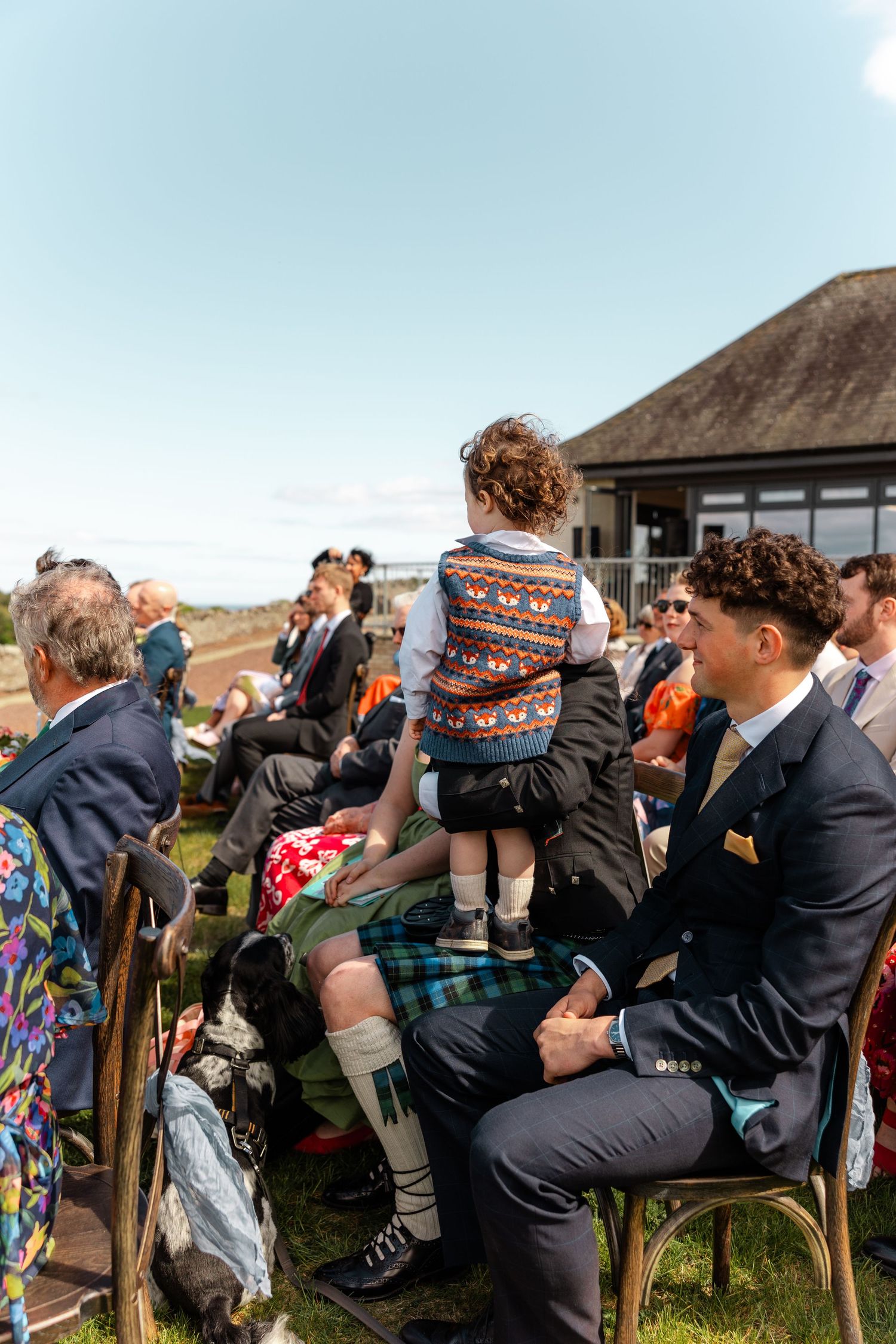 Guests seated outdoors at a wedding ceremony on a sunny day, including a child in a patterned vest sitting on an adult's lap.