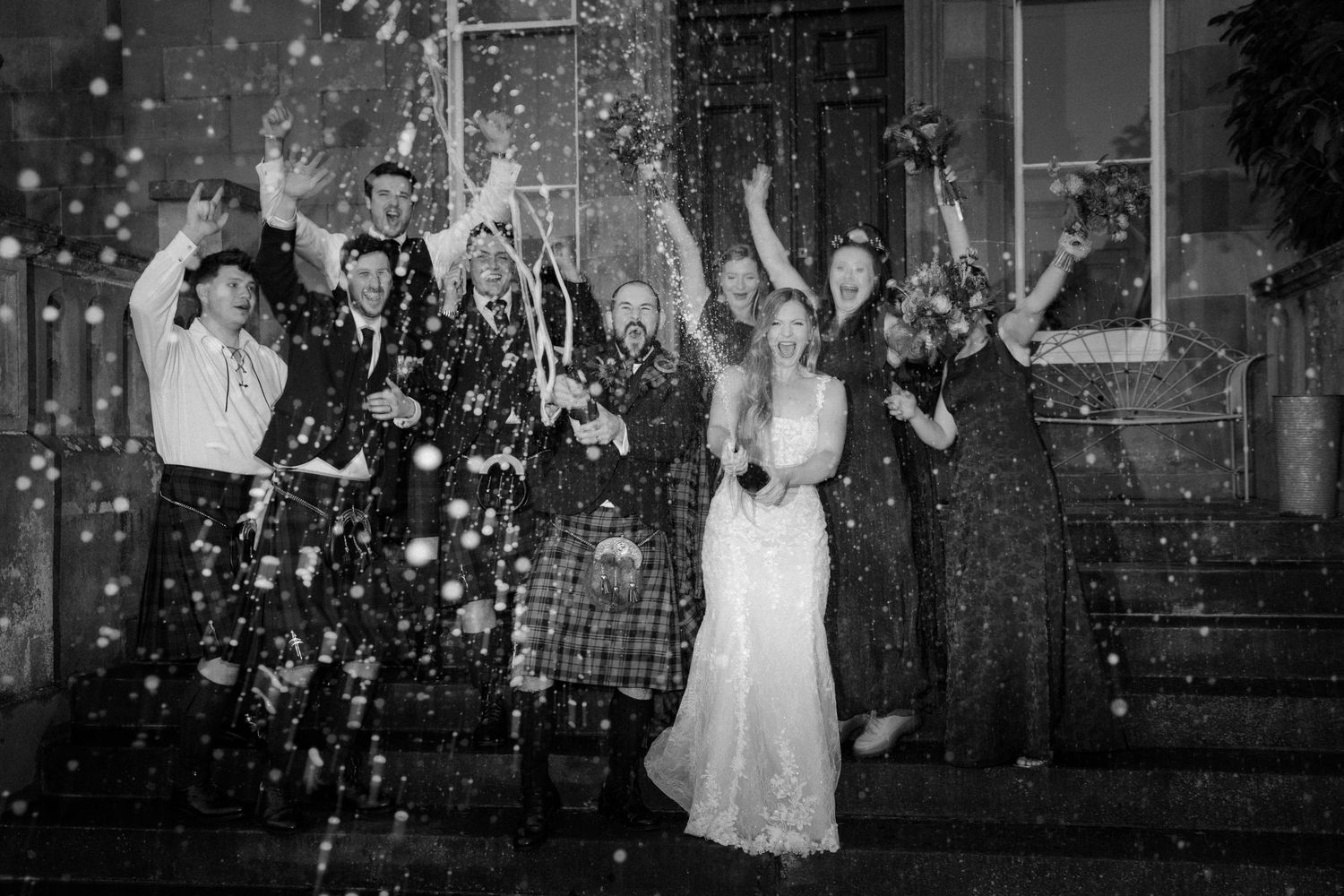 Black and white wedding photo of bride, groom and guests celebrating with confetti on venue steps at night.