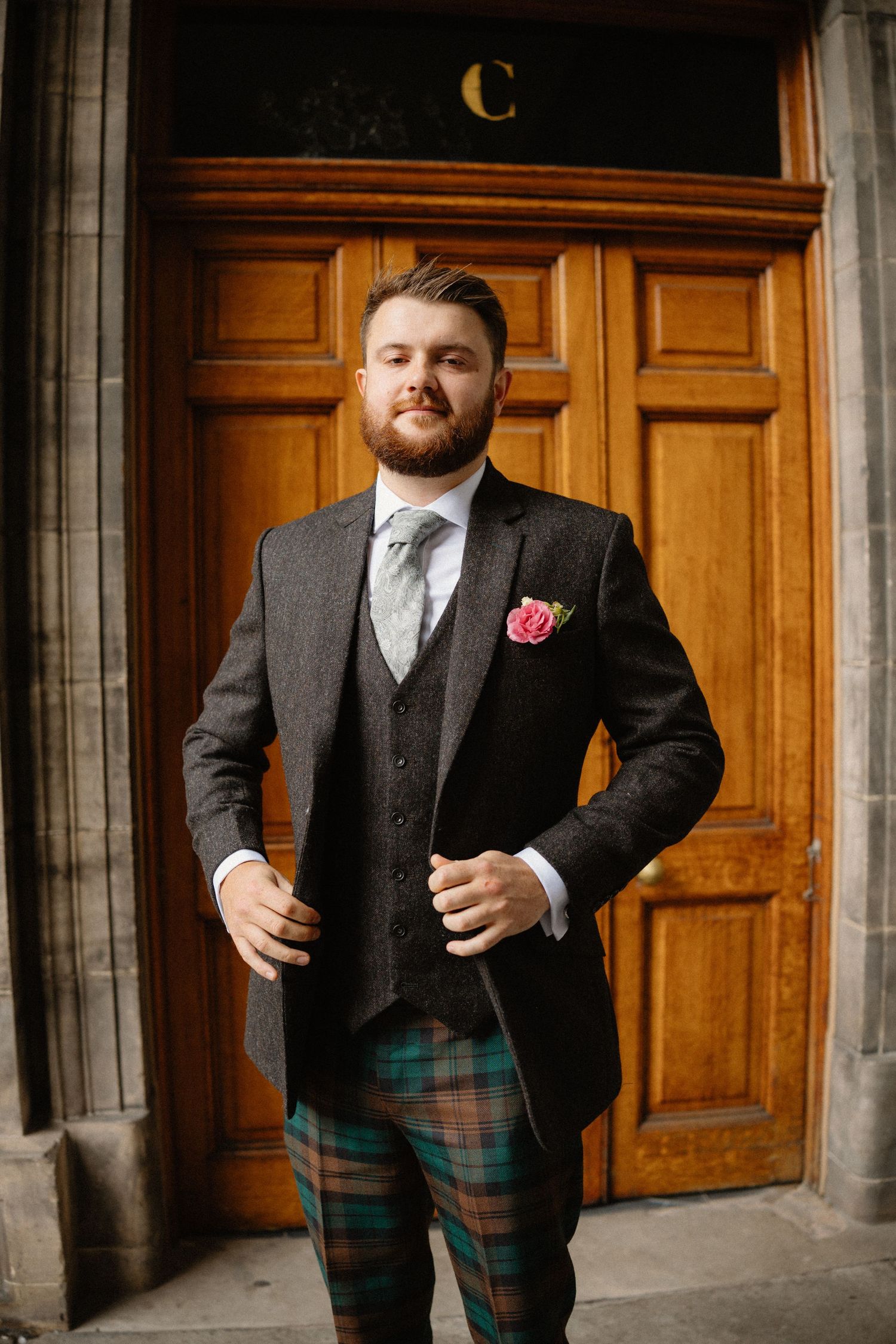 Bearded groom in tartan trousers, grey tweed jacket, and pink boutonniere poses before wooden doors.