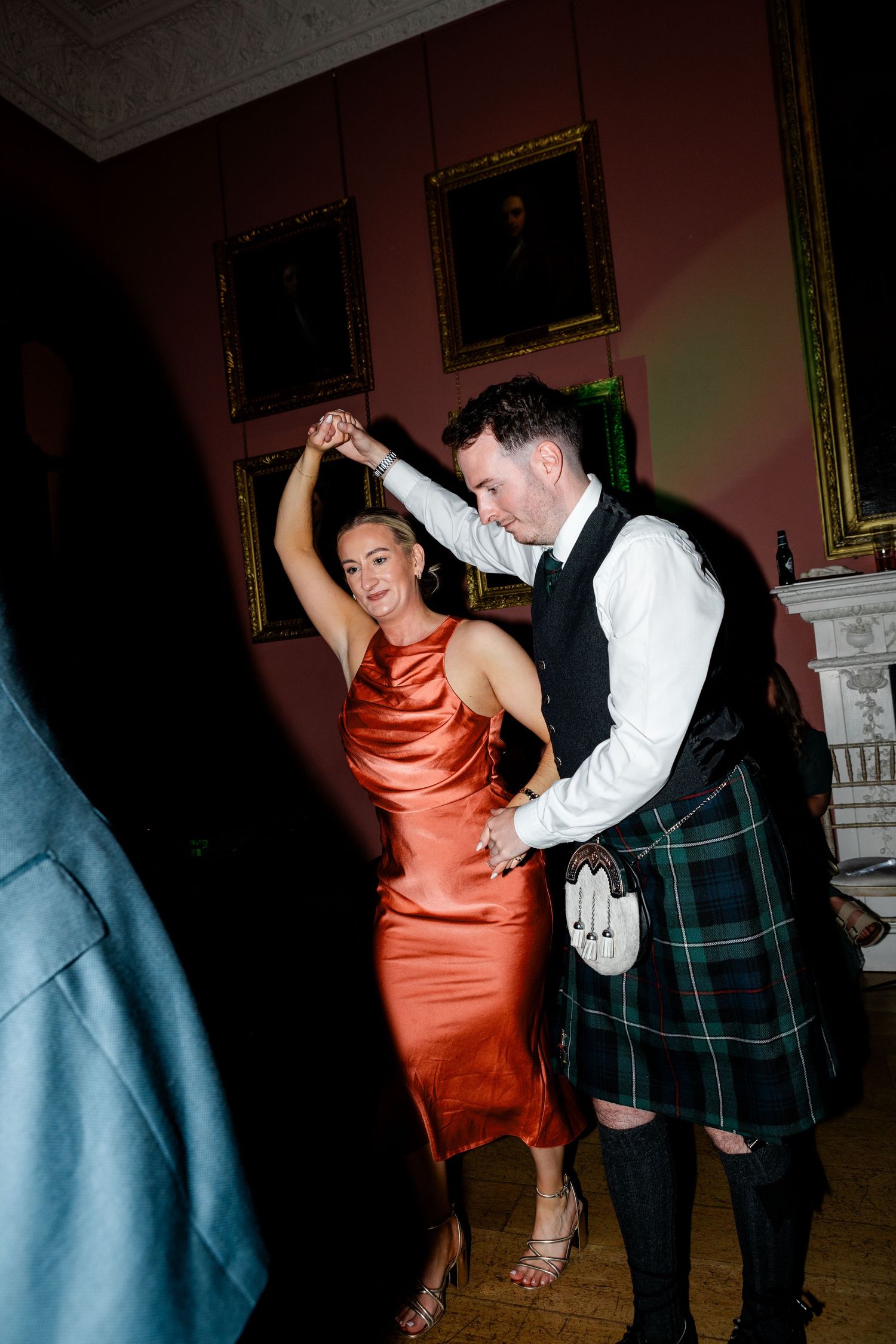 Woman in orange satin dress dancing with man in Scottish tartan kilt at wedding reception.
