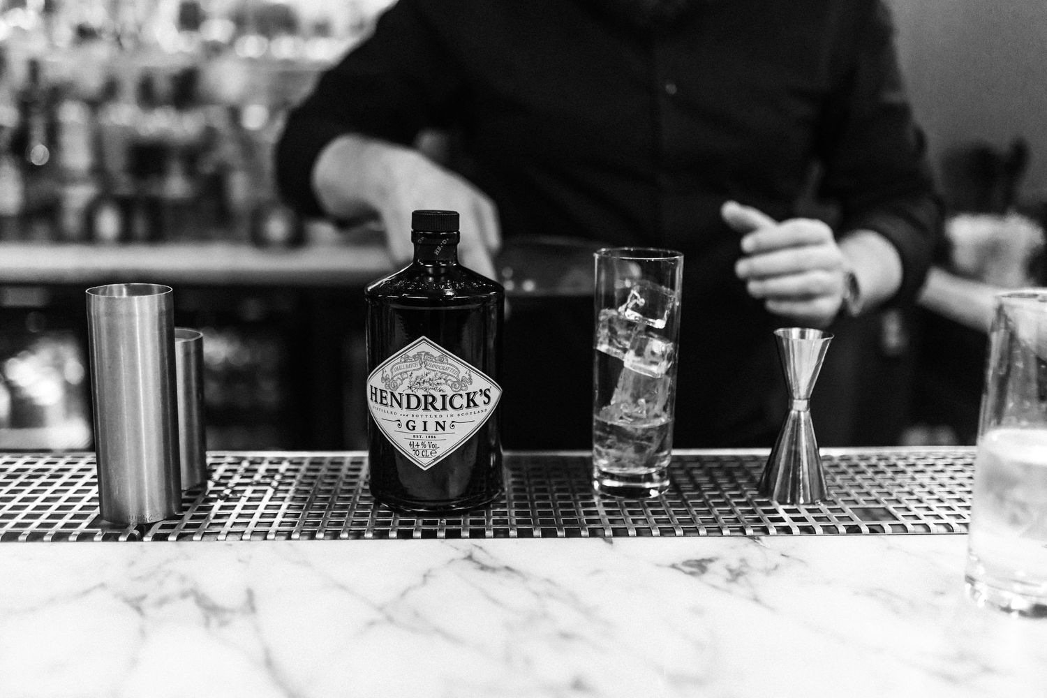 A bottle of Hendricks Gin on a marble bar counter with a bartender mixing drinks behind.