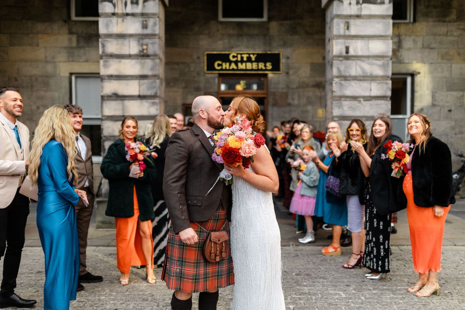 Newlyweds kissing outside City Chambers as guests throw confetti in a colorful wedding celebration.