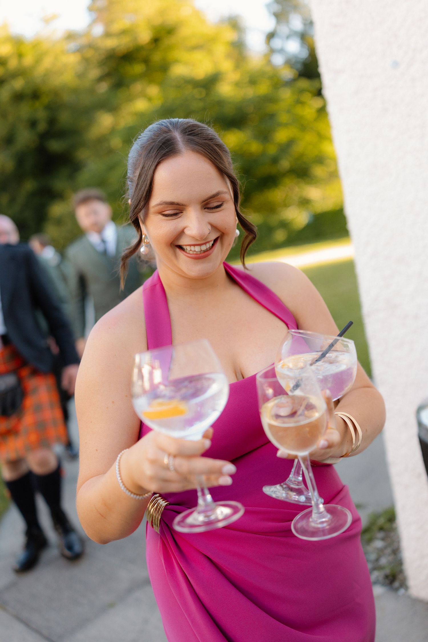 Smiling bridesmaid in pink halter dress holding three gin cocktail glasses at an outdoor wedding reception.