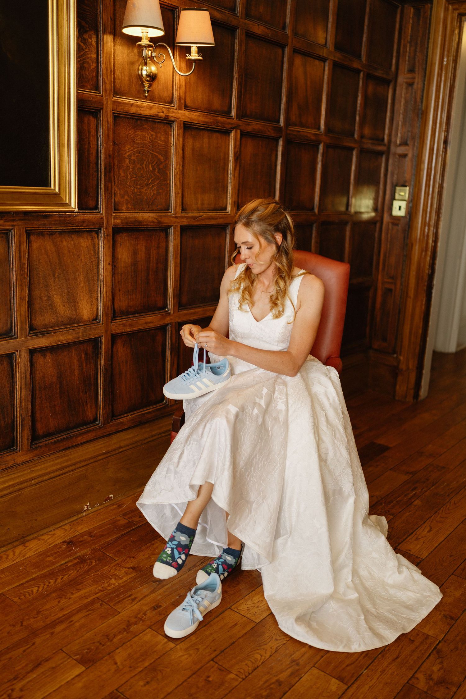 Bride in white wedding gown sits in leather chair holding blue sneakers wearing colorful socks in wood-paneled room.