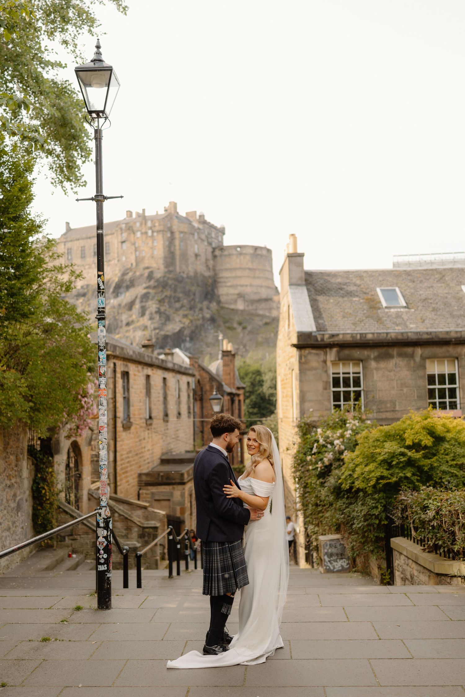 Bride and groom embrace on cobblestone steps in Edinburgh with historic castle on rocky hill backdrop.