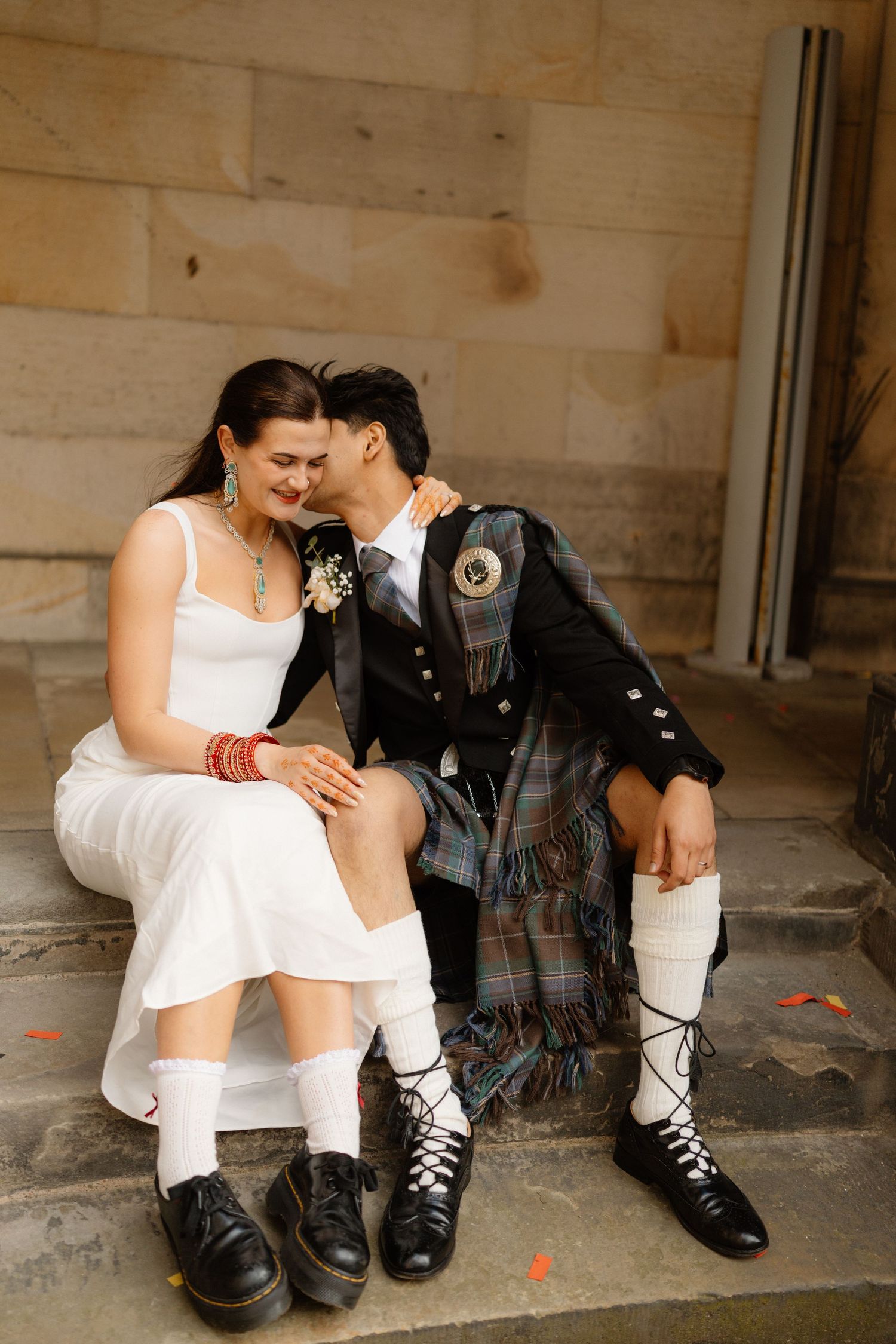 Bride and groom sitting on stone steps, she in white dress with Doc Martens, he in Scottish Highland kilt attire.