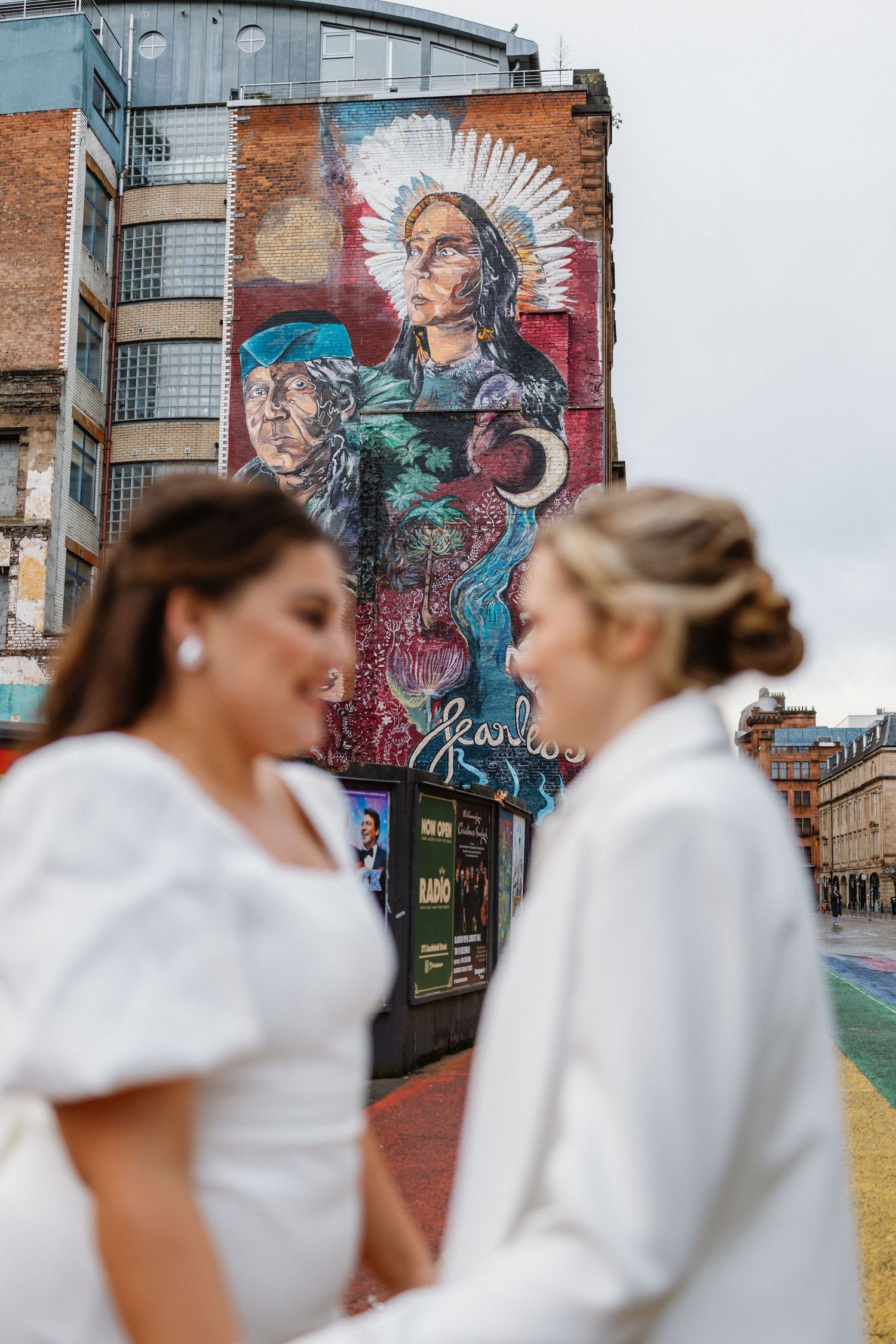 Two brides in white stand before a large vibrant street art mural on a city building.