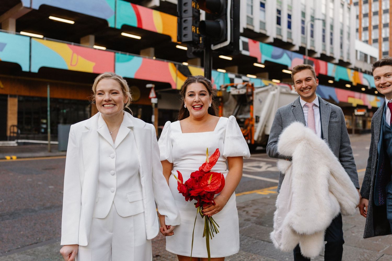 Two brides in white outfits smile holding red roses on a city street, with guests walking behind them.
