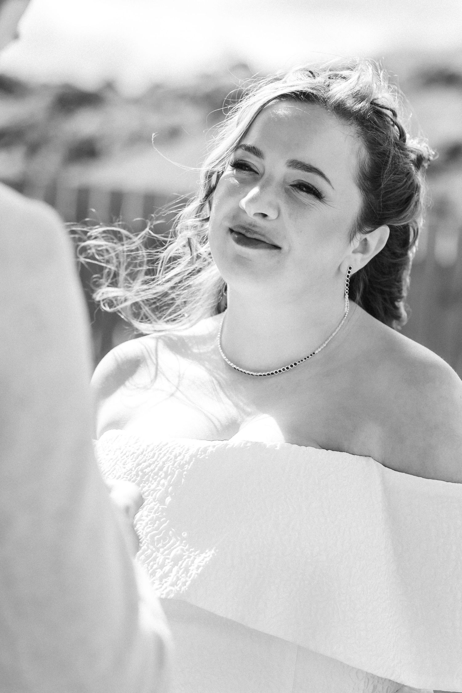 Black and white portrait of smiling bride in off-shoulder gown with hair blowing in wind.