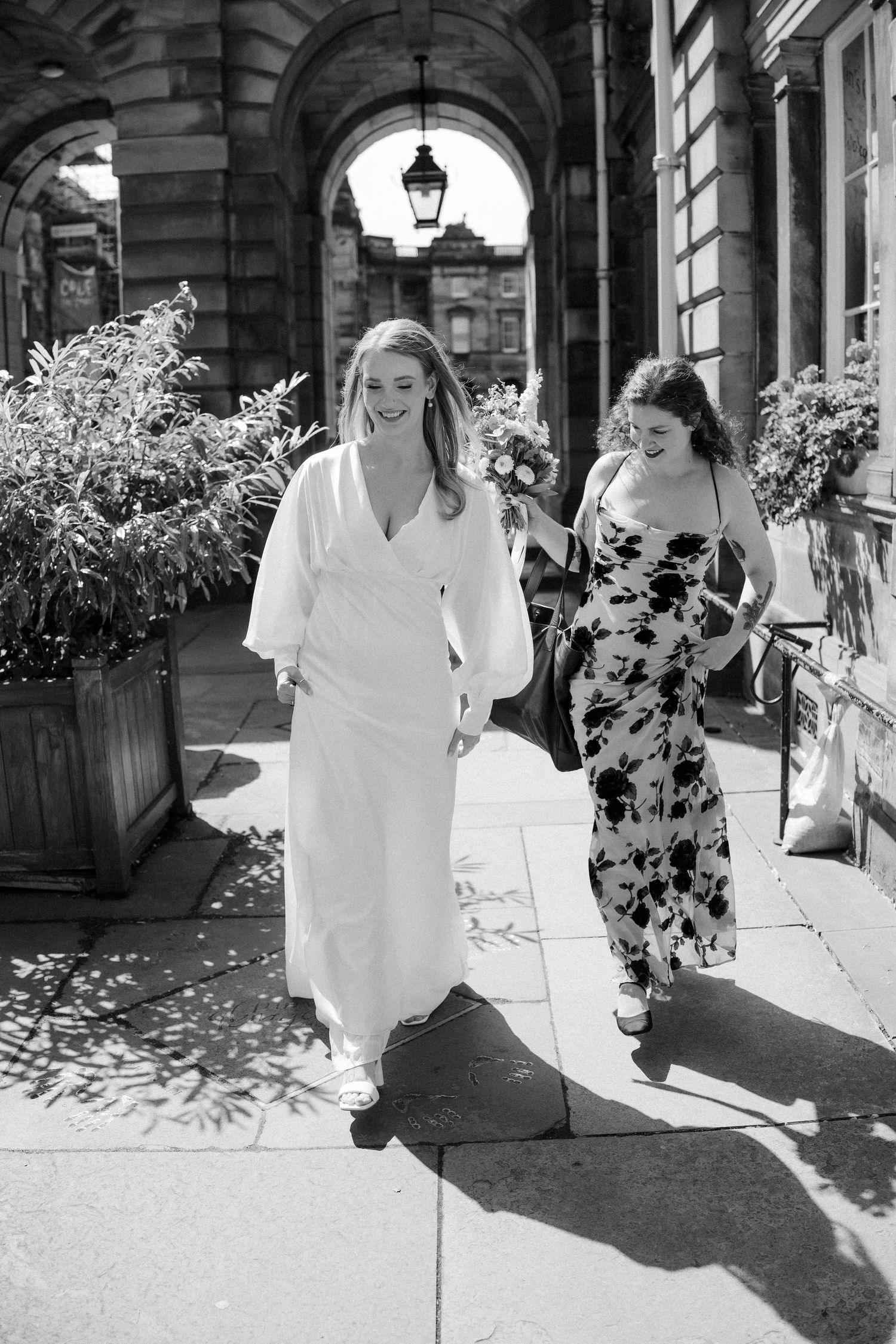 Bride in elegant white dress walks with bridesmaid along sunlit historic Edinburgh street.