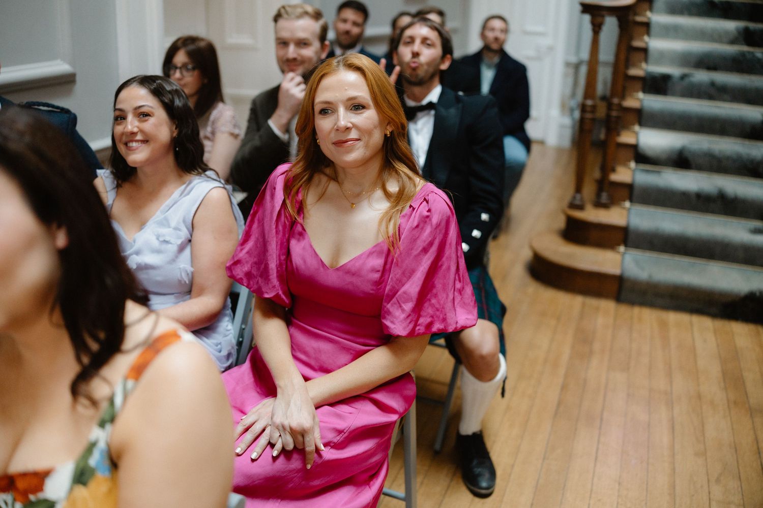 Wedding guest in a bright pink dress smiles warmly while seated during the ceremony.