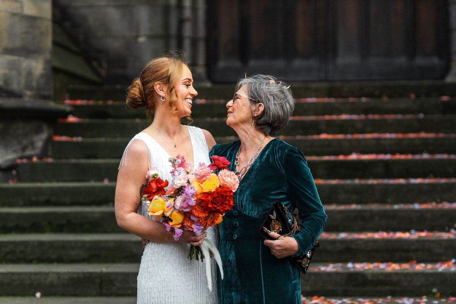 Bride in white gown holding colorful bouquet smiling with older woman in teal dress on stone steps.