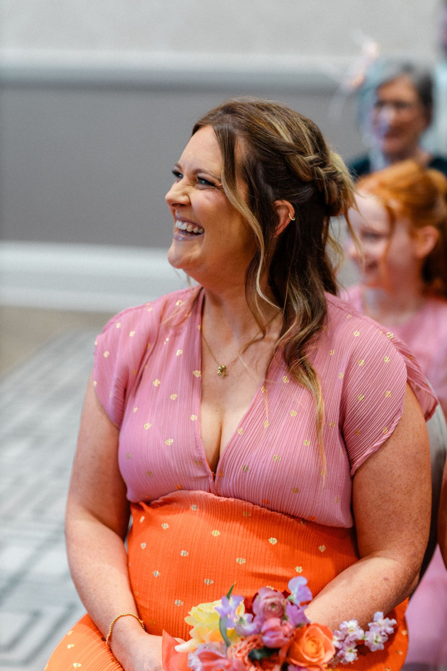 Smiling bridesmaid in pink and orange dress holding a colorful bouquet at a wedding ceremony.