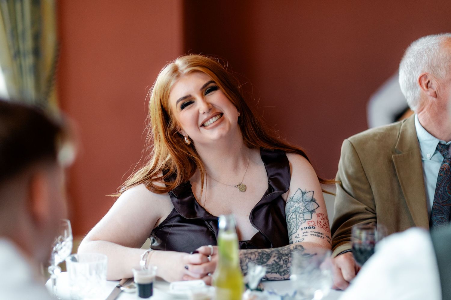 Smiling redhead woman with tattoos in black dress sits at elegantly set wedding reception dinner table.