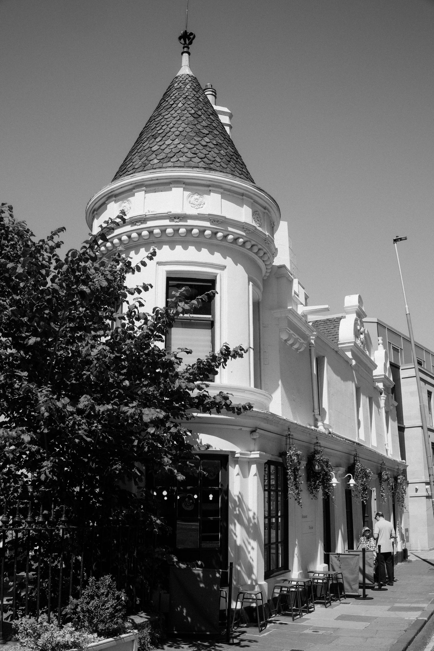 Black and white photo of Victorian building with conical turret tower and outdoor cafe seating.