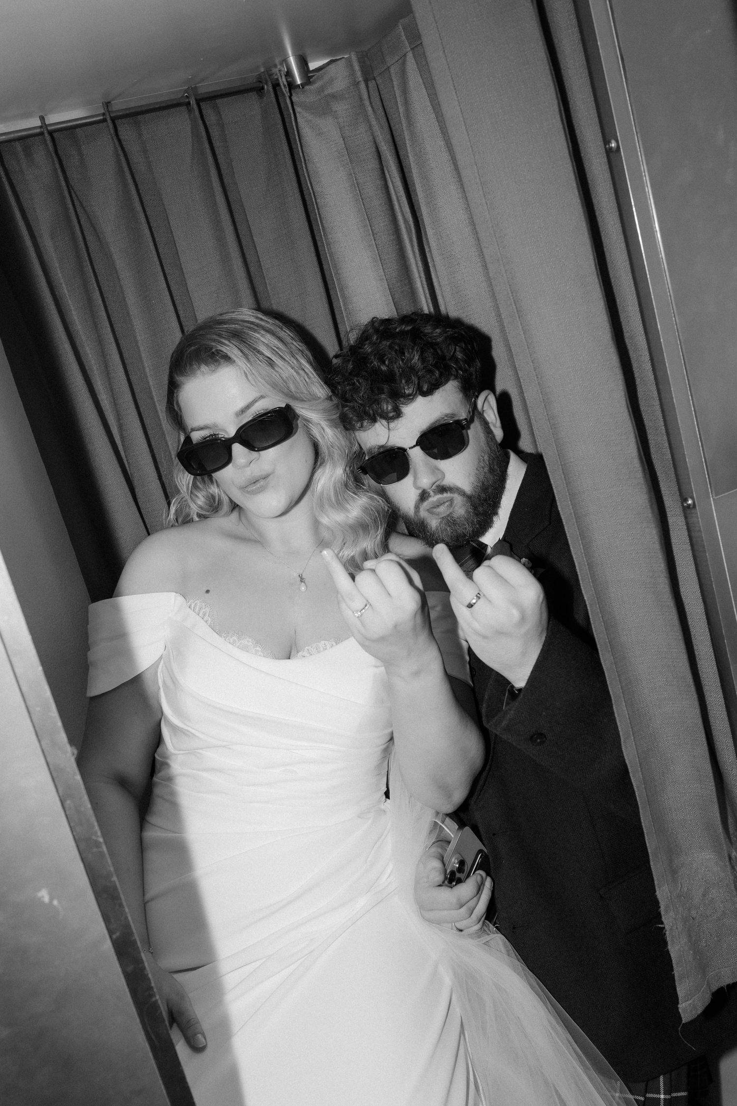 Bride and groom wearing sunglasses pose playfully in photo booth at wedding reception in black and white.