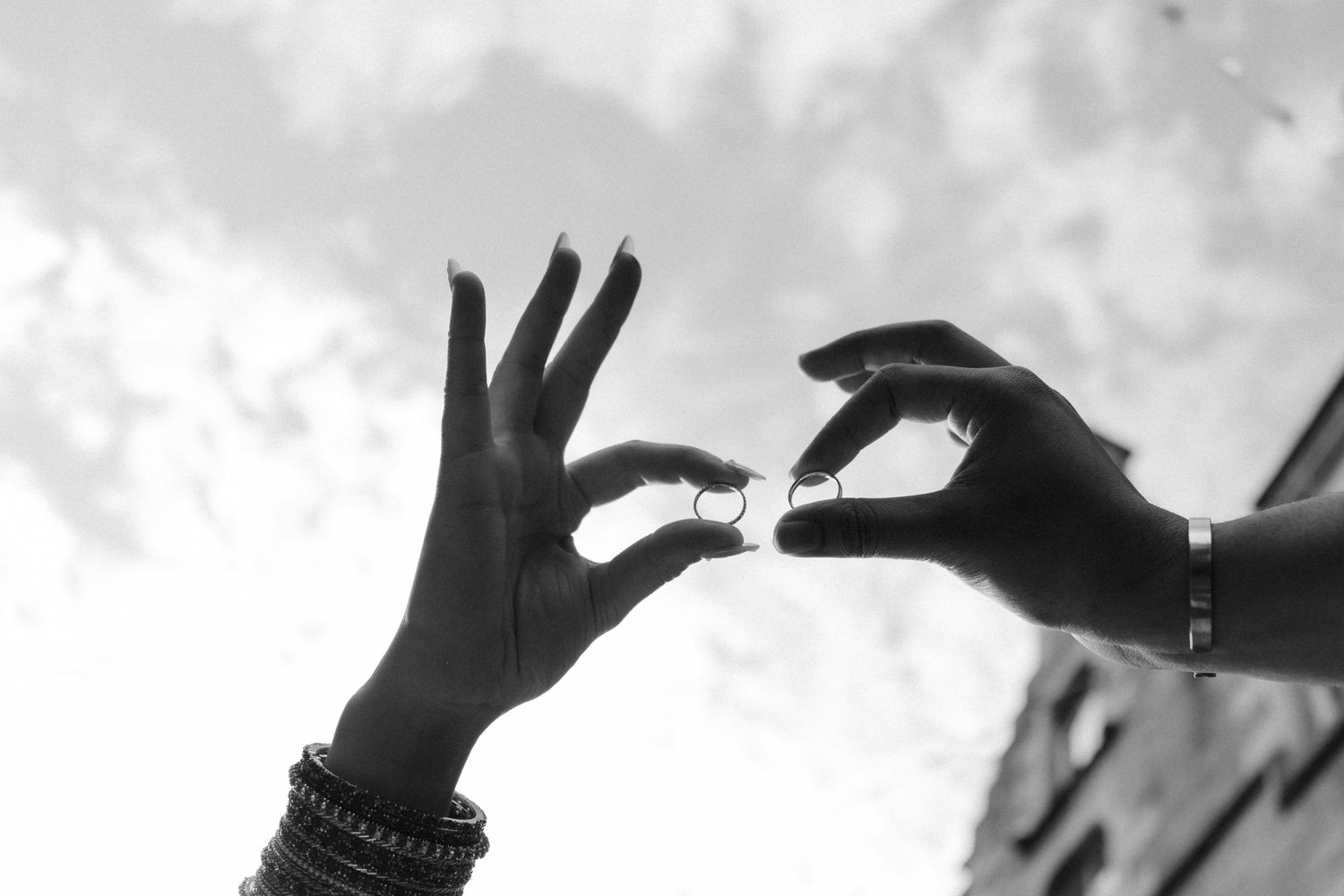 Black and white photo of two hands reaching toward each other, exchanging rings against a cloudy sky.