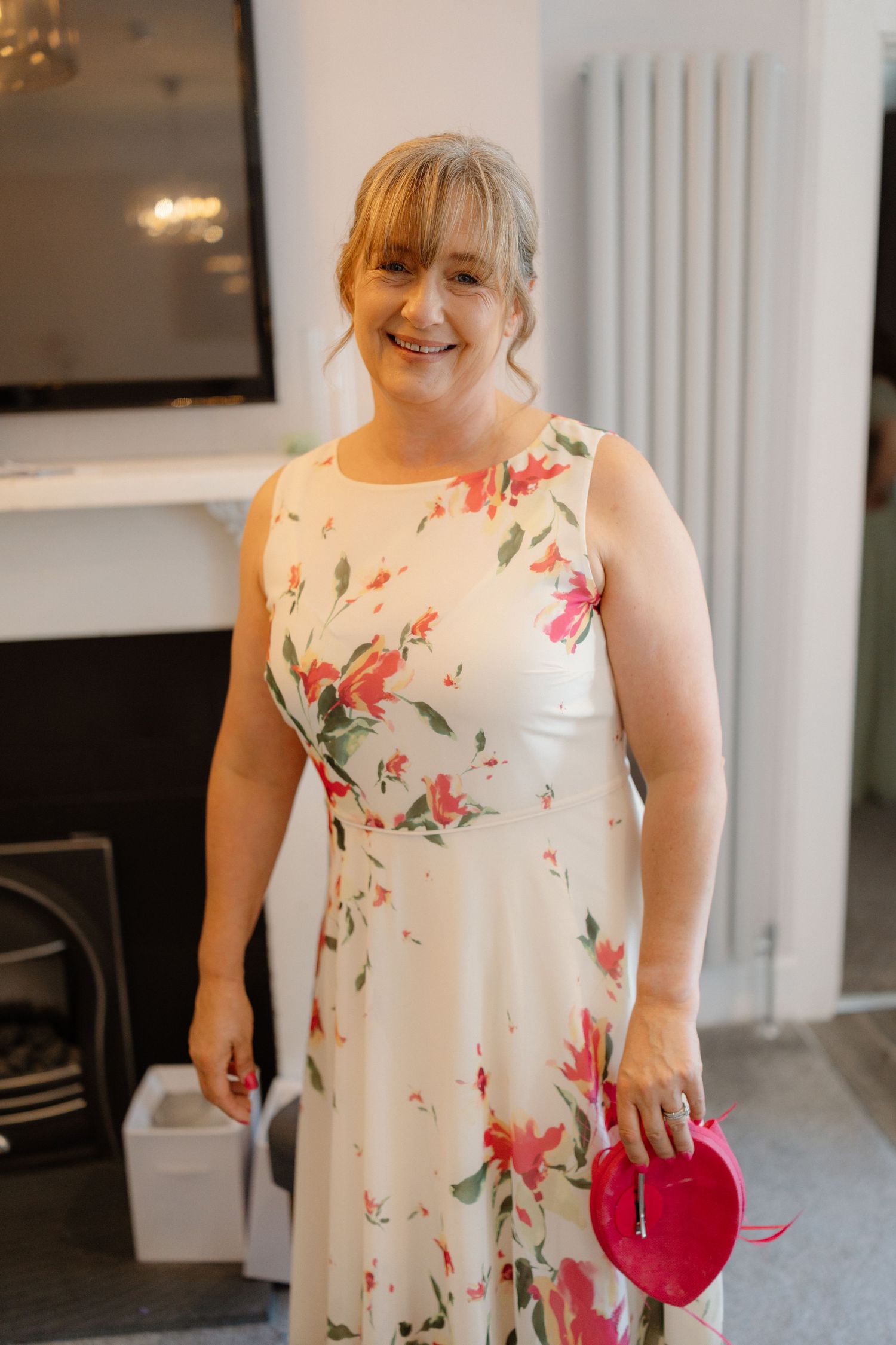 Woman smiling in white floral dress holding red heart clutch bag at a wedding event.