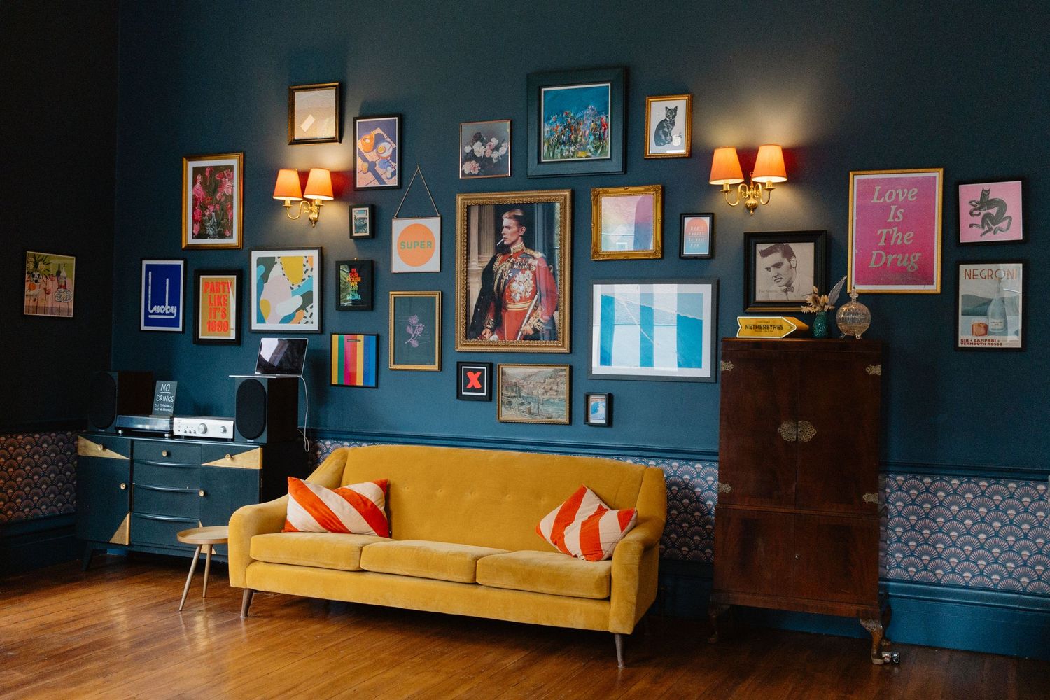 A mid-century modern living room with teal walls, yellow velvet sofa, and gallery wall of framed artwork and photographs.