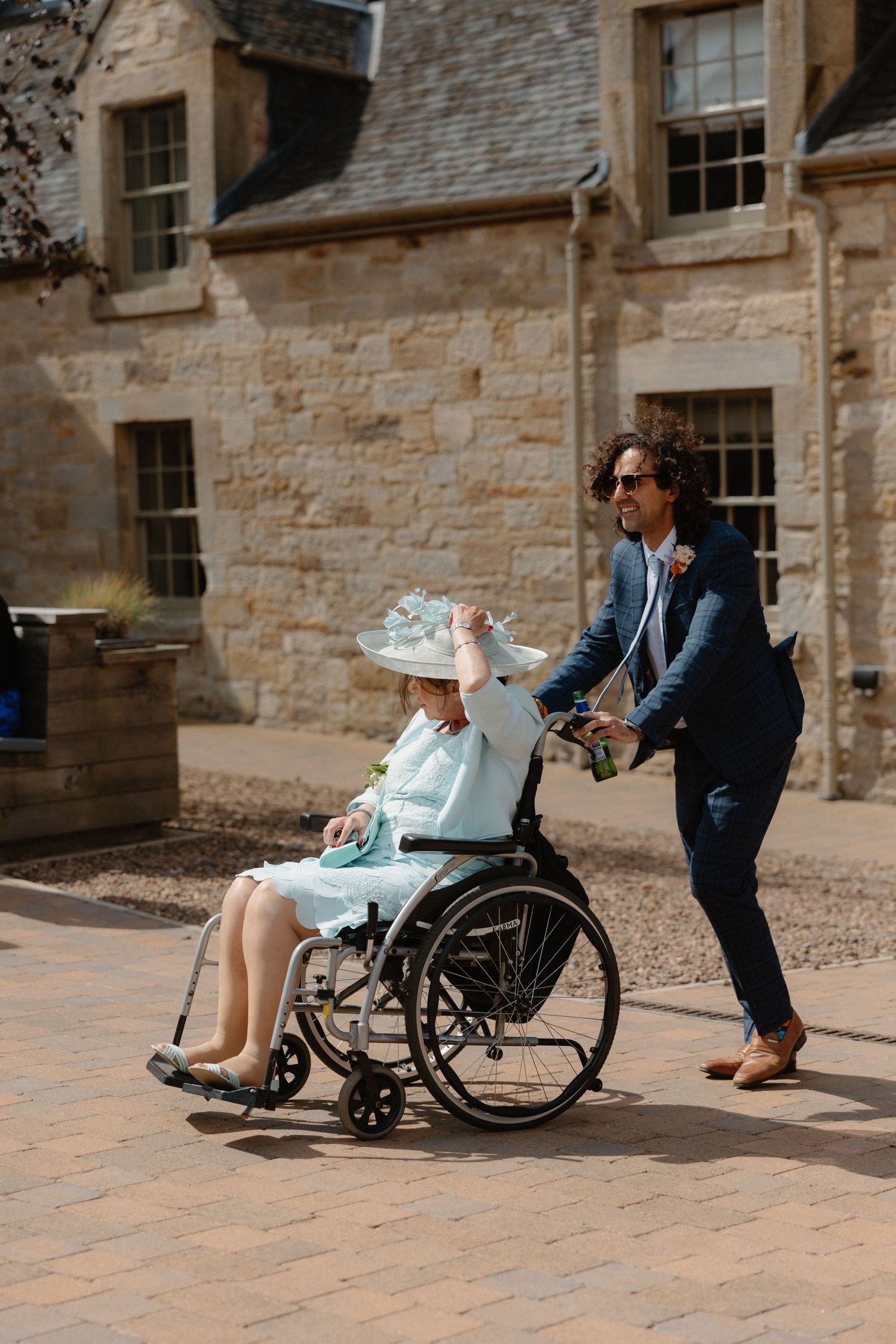 Man in navy suit pushing a woman in a wheelchair wearing a blue hat outside a stone building on a sunny day.