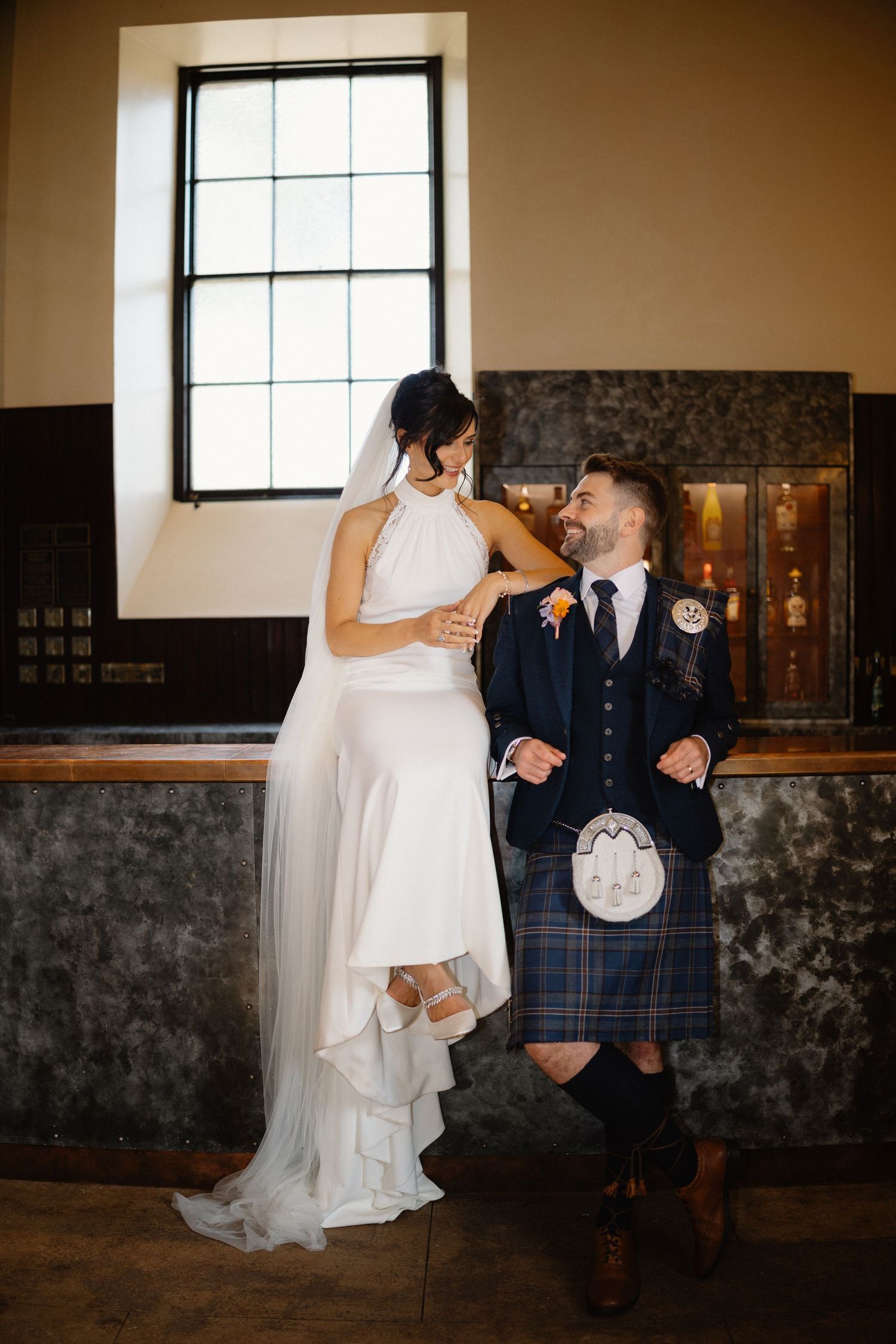 Bride and groom sharing a joyful moment indoors, groom wearing a blue Scottish kilt beside a large window.