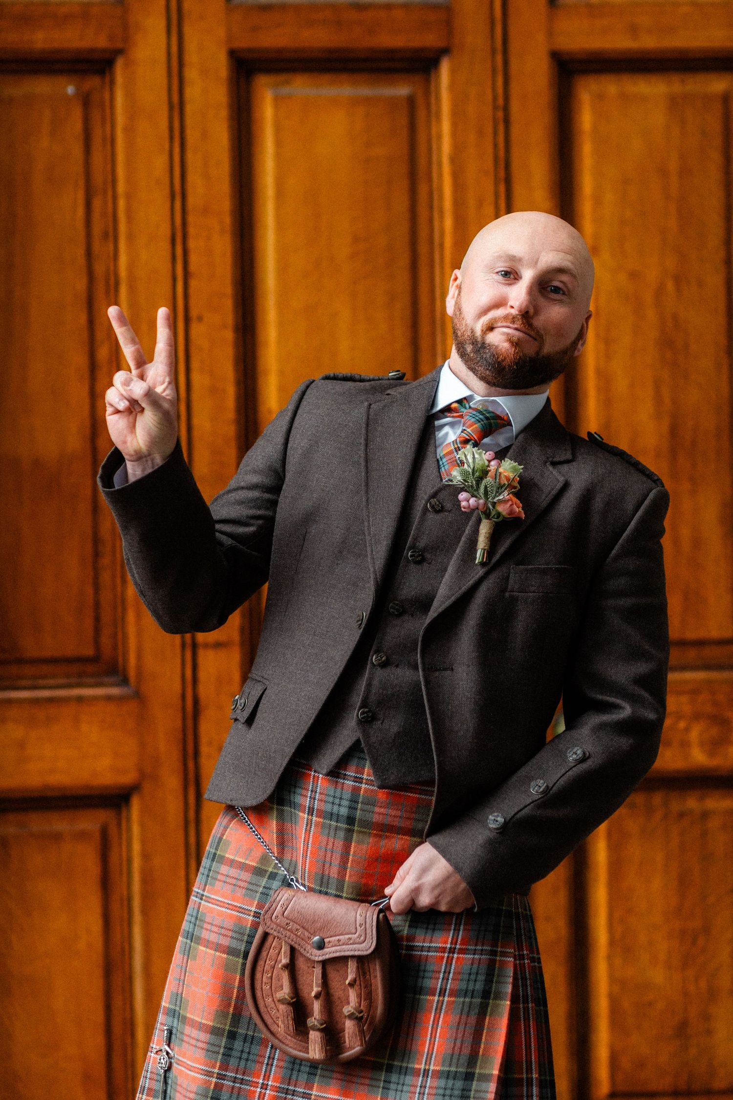 Bald bearded man in traditional Scottish kilt outfit with tartan sporran and floral boutonniere making peace sign.