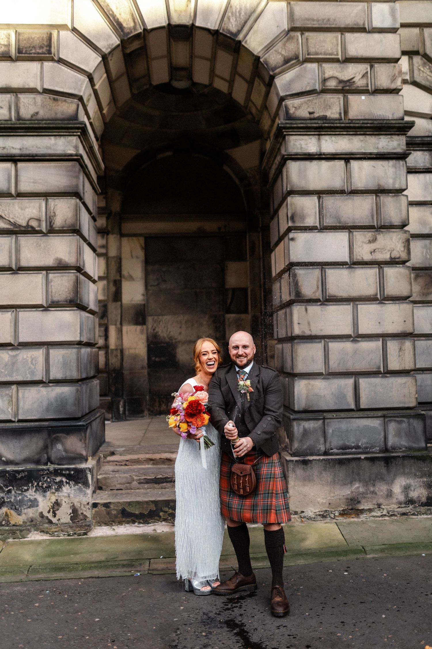 A laughing bride holding colorful flowers stands with a groom in a tartan kilt outdoors.