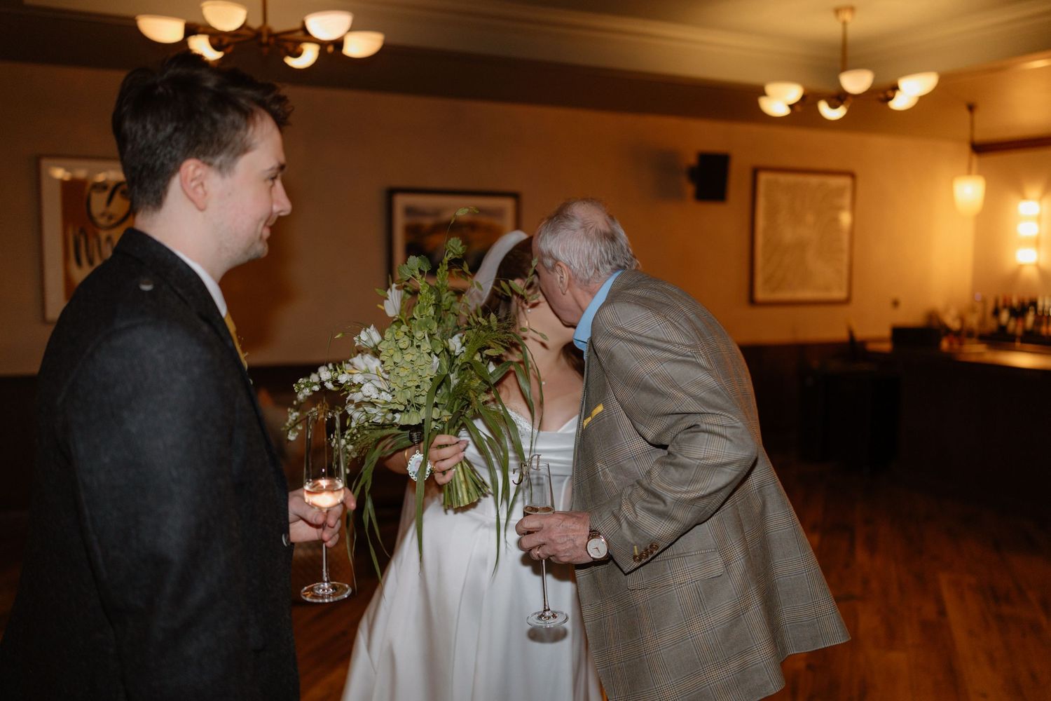 An older gentleman kisses a bride on the cheek as a groom stands nearby holding champagne at a wedding reception.