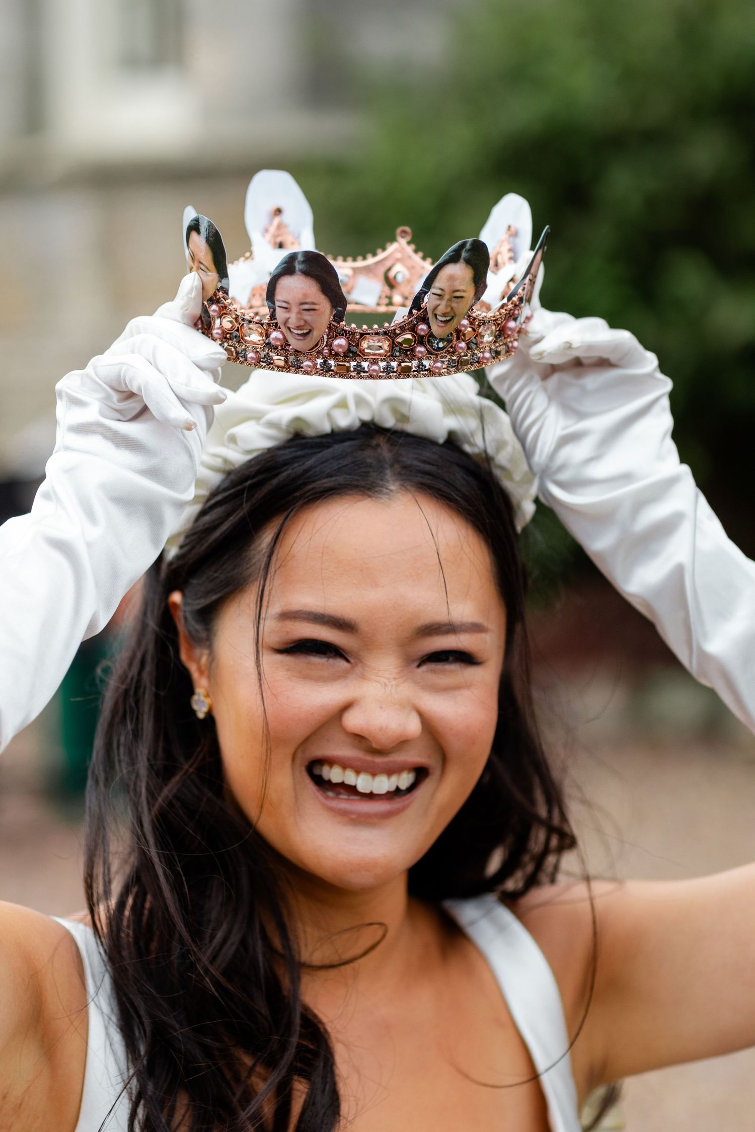 Joyful bride wearing decorative crown with photos, white gloves, and wedding dress, laughing outdoors at Scottish wedding.