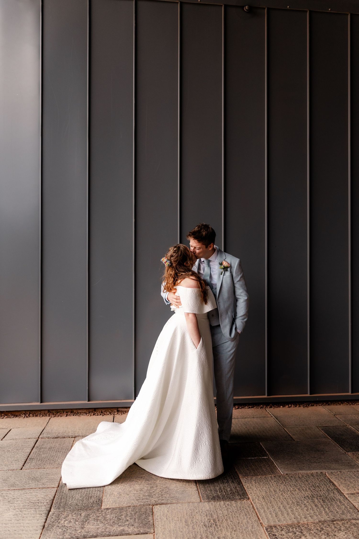 Bride and groom share a kiss against a modern grey panelled wall in wedding attire.