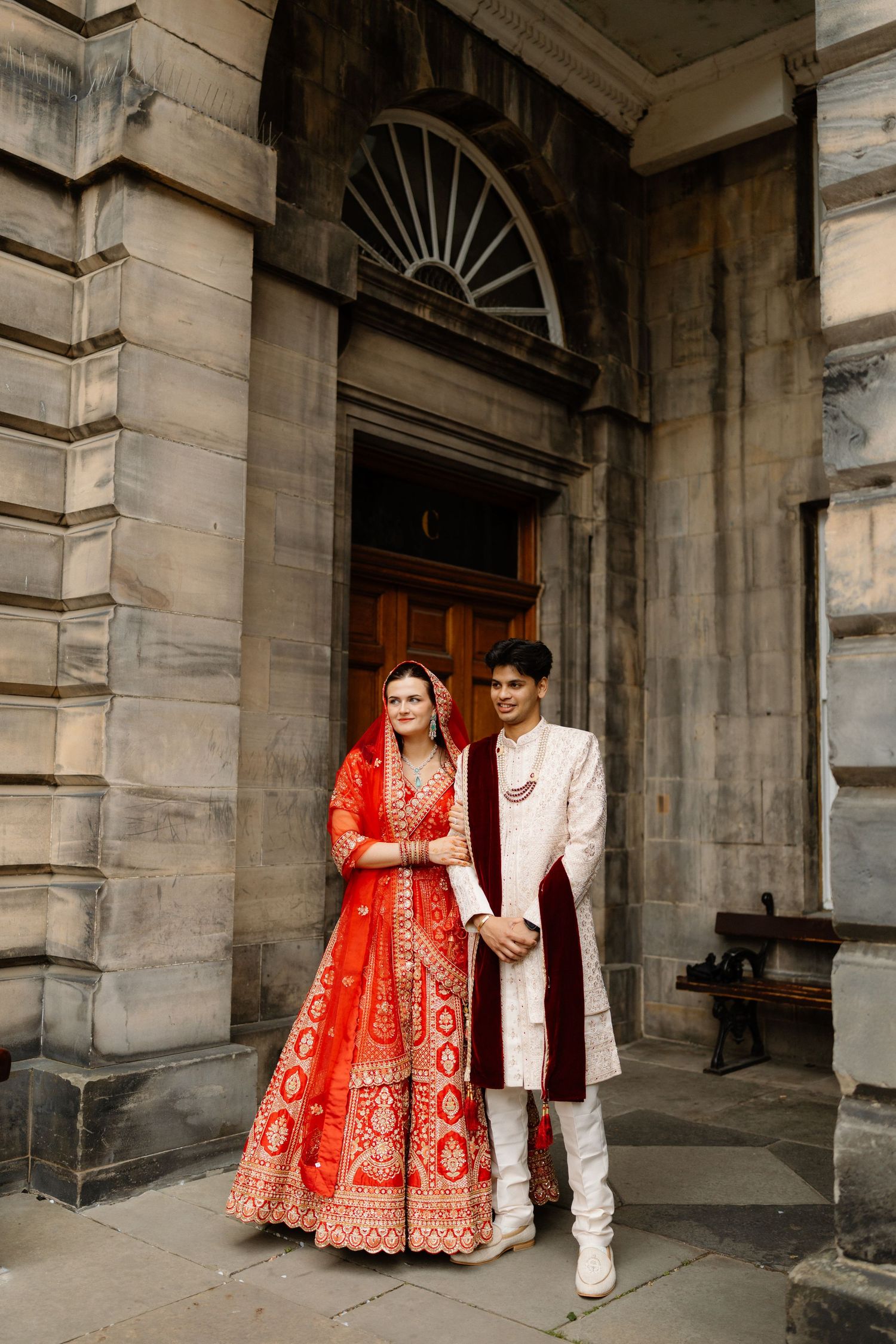 Indian bride in red lehenga and groom in white sherwani pose at historic Edinburgh building entrance.