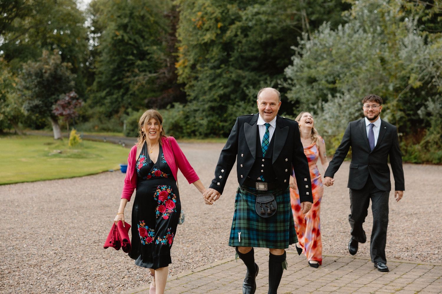 Wedding guests walking along gravel path, man in blue jacket and green tartan kilt leading group.