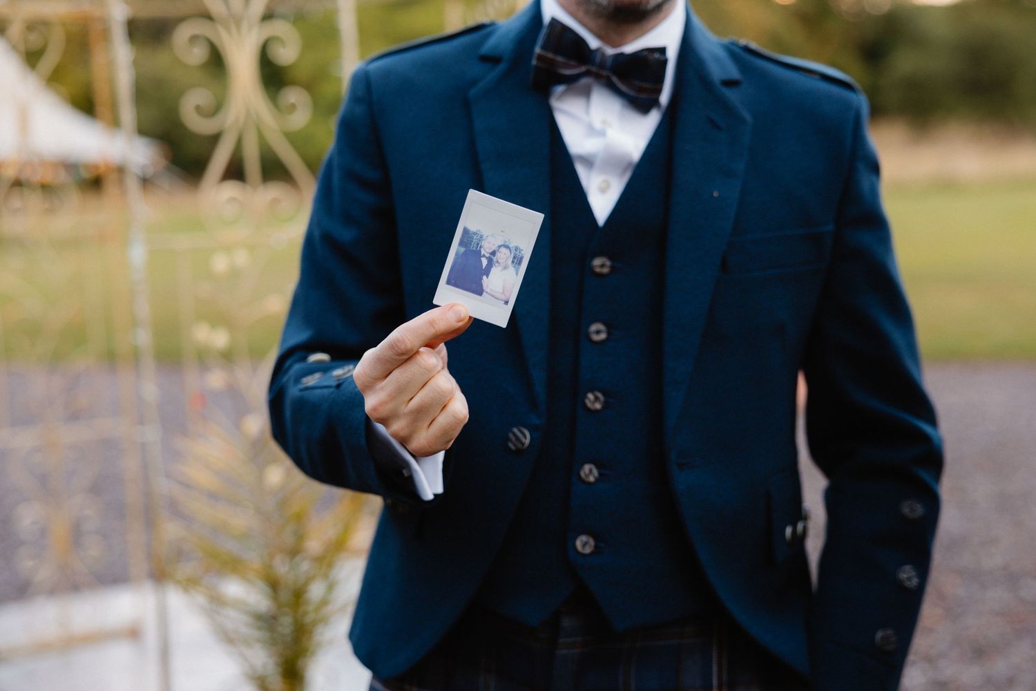 Groom in navy suit and bow tie holding a Polaroid wedding photo outdoors.