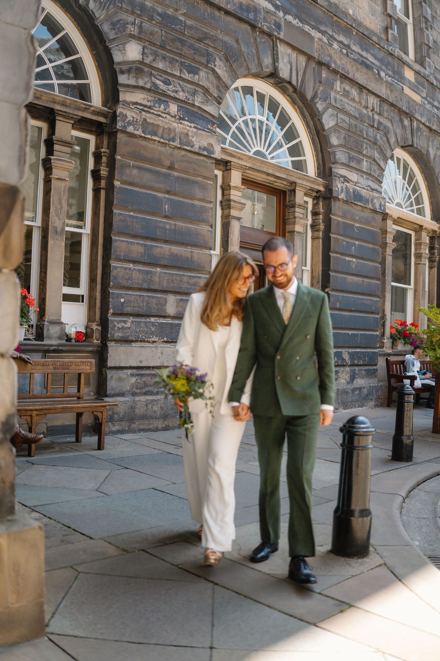 Newlywed couple in white pantsuit and green suit walking hand in hand outside Georgian building.