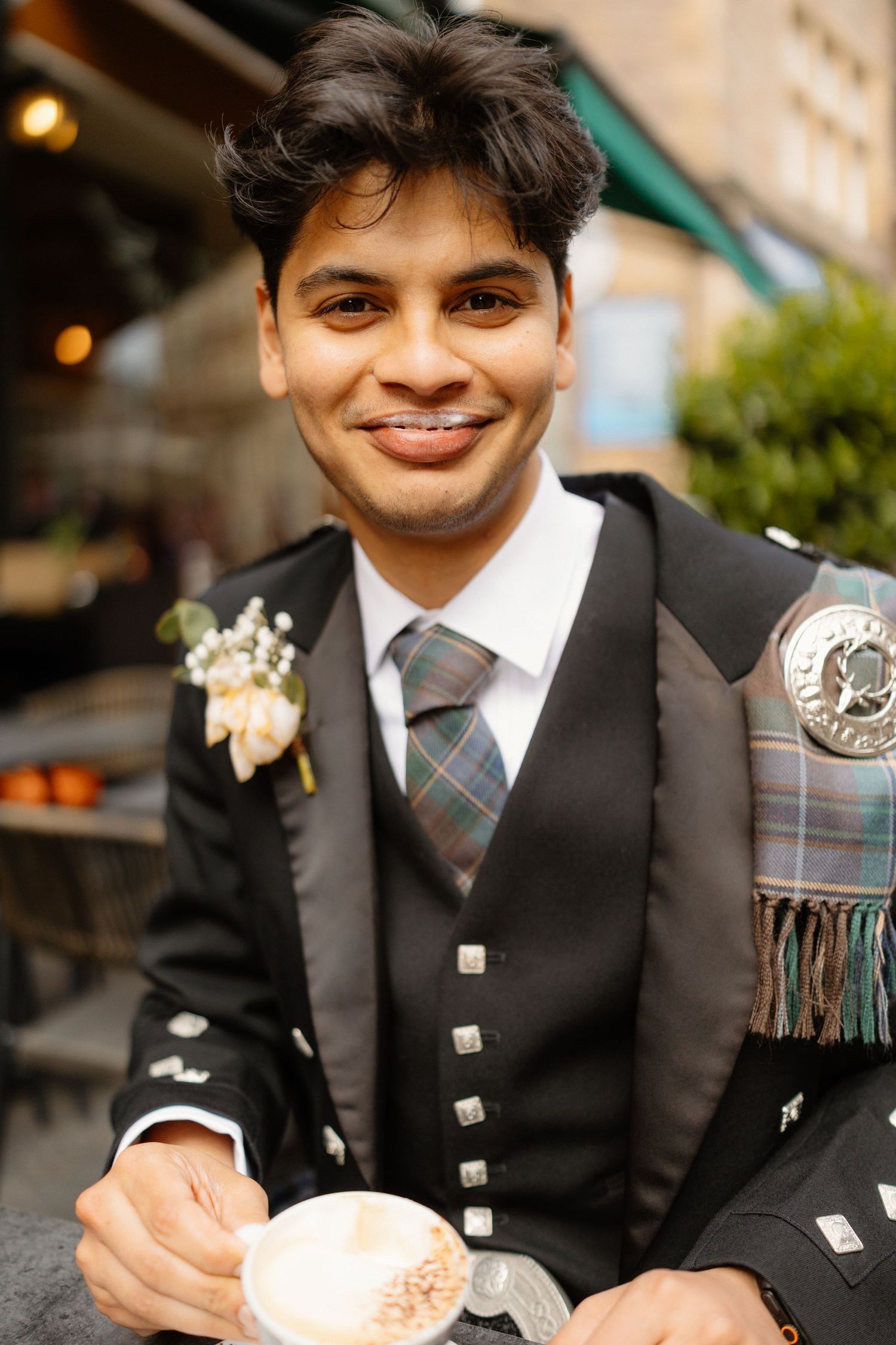 Smiling groom in traditional Scottish Highland dress with tartan kilt and boutonniere holding a latte at outdoor cafe.
