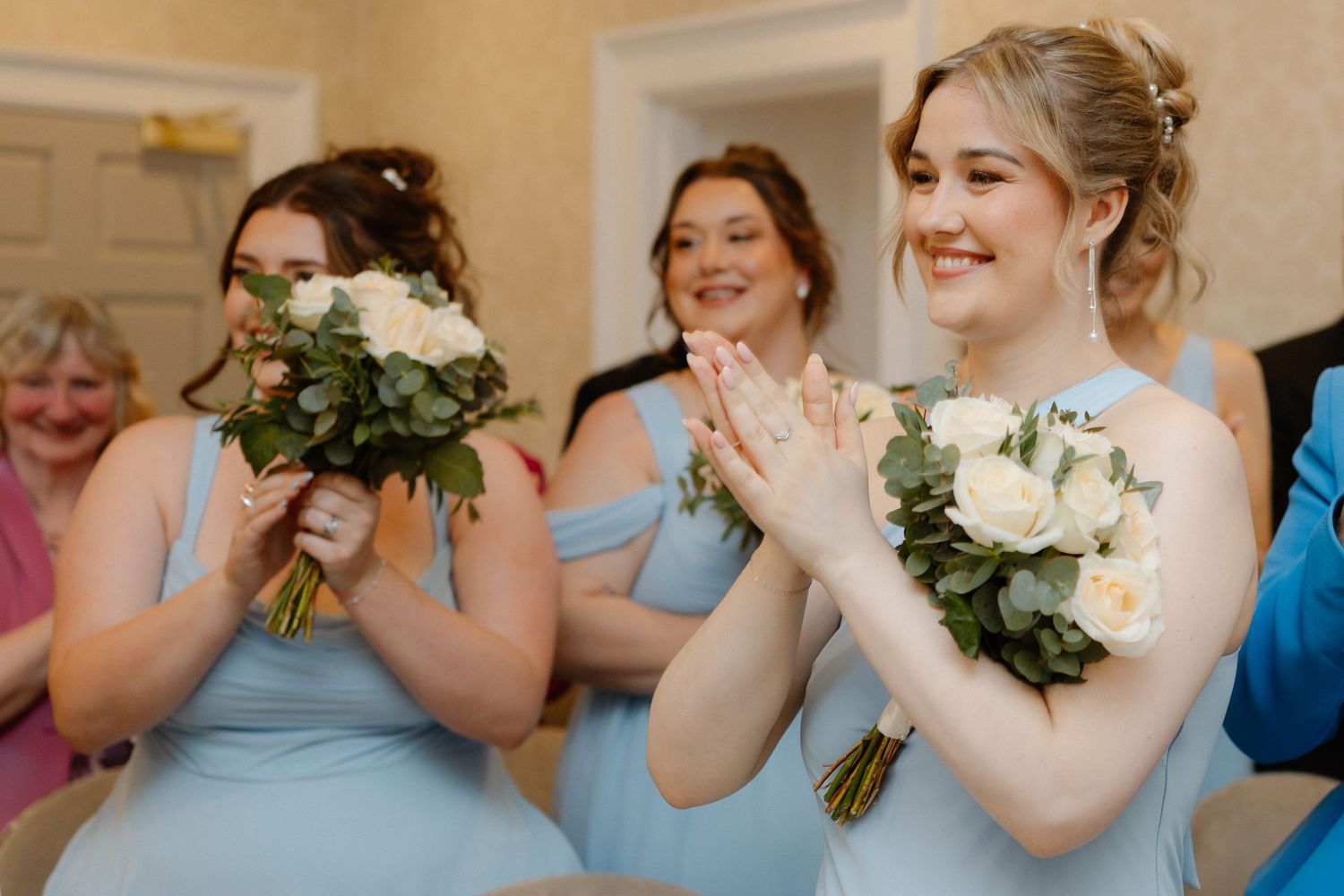 Bridesmaids in light blue dresses clapping and holding white floral bouquets during a wedding ceremony.