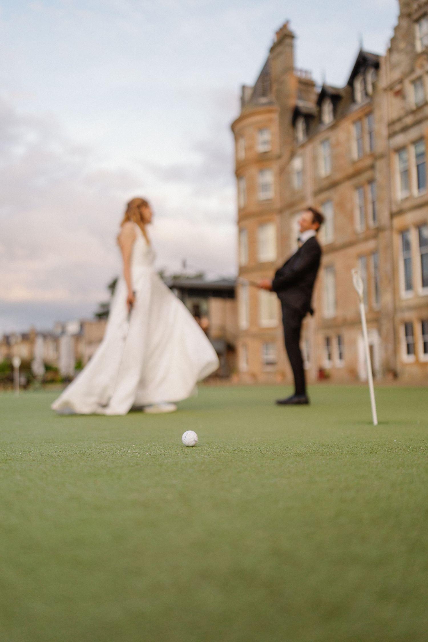 Bride and groom posing on golf course green with golf ball in foreground at grand hotel.