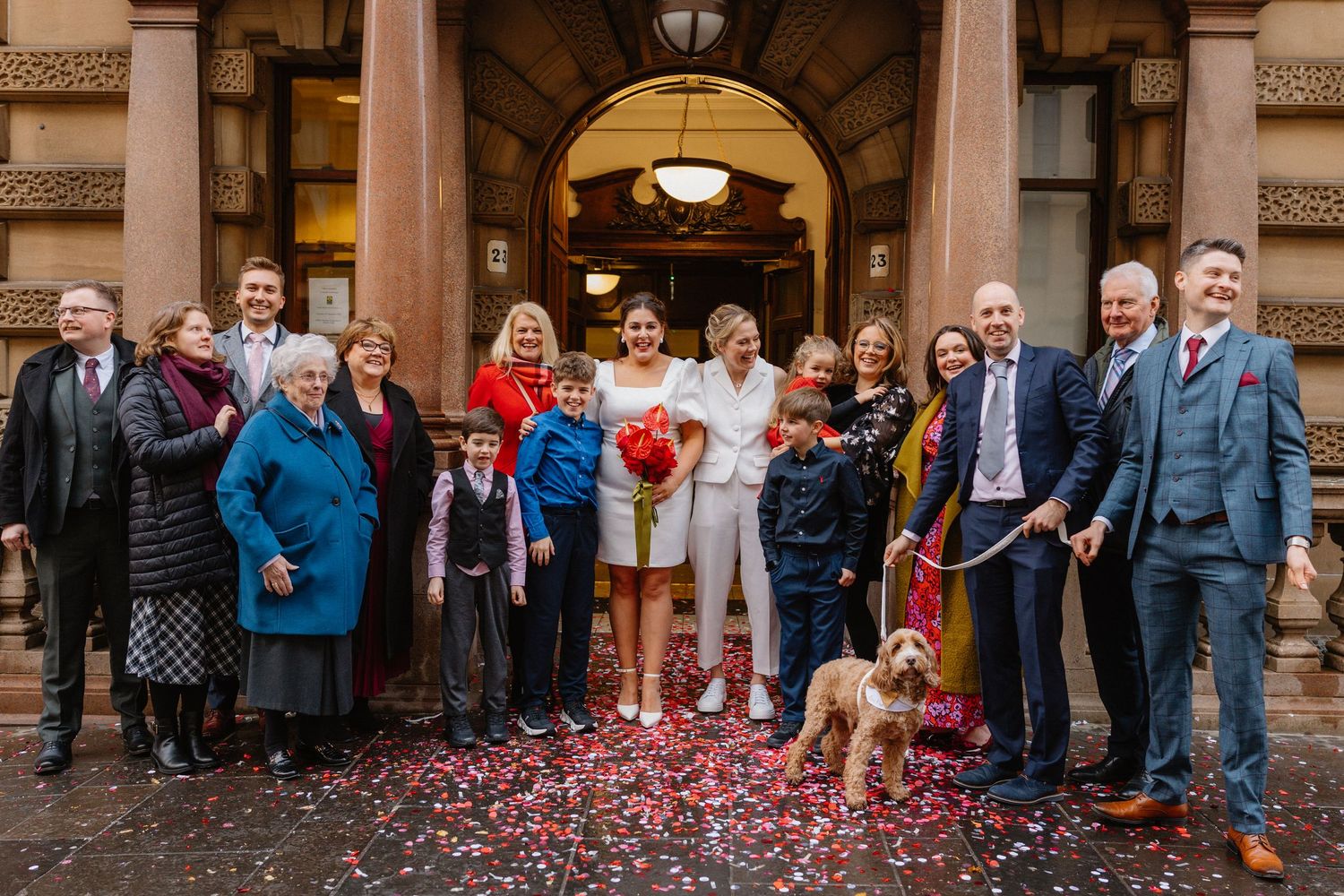 Two brides in white celebrate with family and a dog outside a venue covered in red confetti.