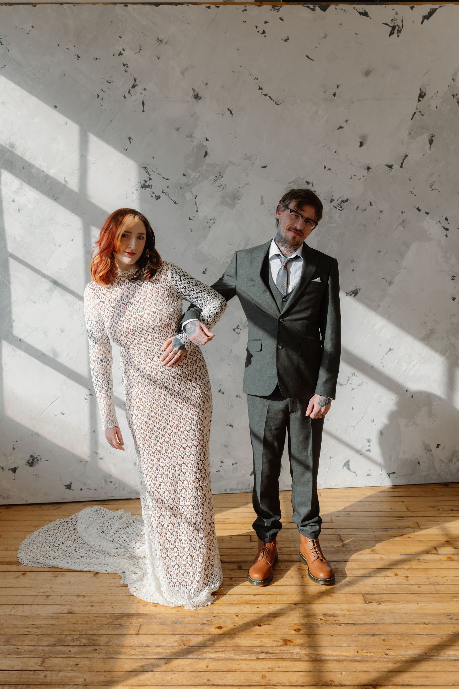 A bride in a white lace gown and groom in a dark suit pose elegantly in a sunlit industrial loft.