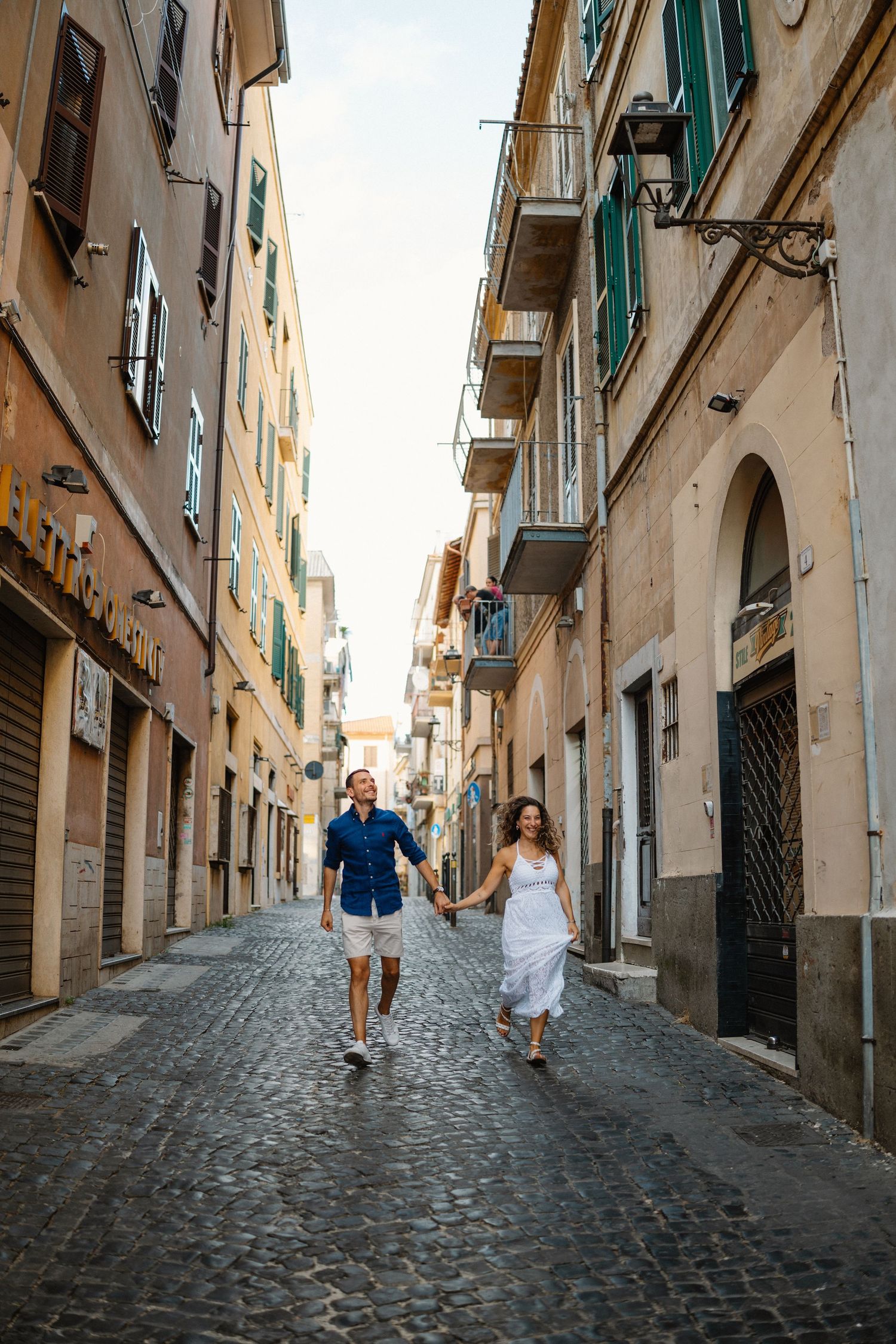 Couple walking down a charming cobblestone street in an old European town with white buildings and green shutters.