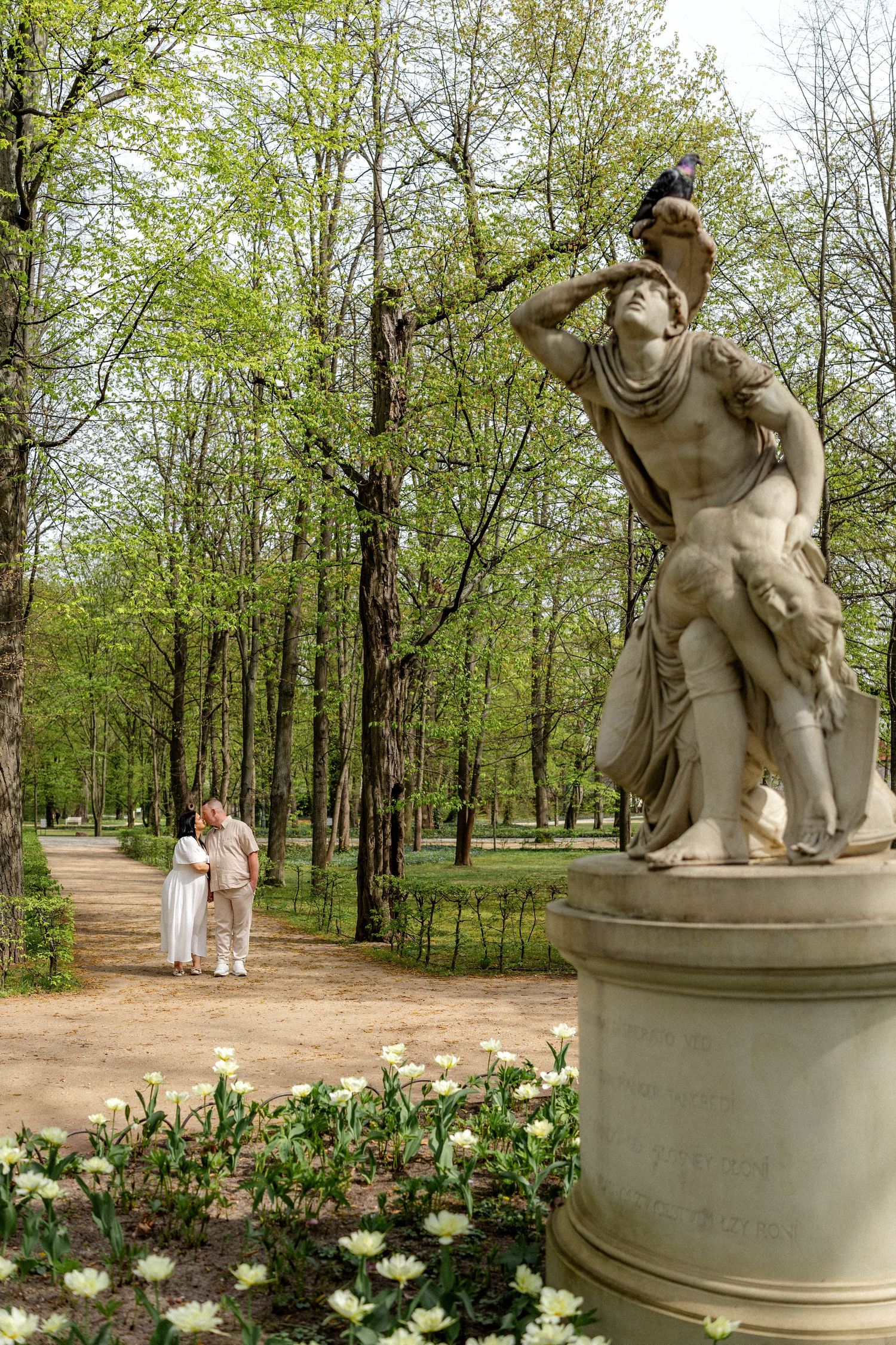 Scenic garden path with classical stone statue and blooming flowers in a serene park setting.